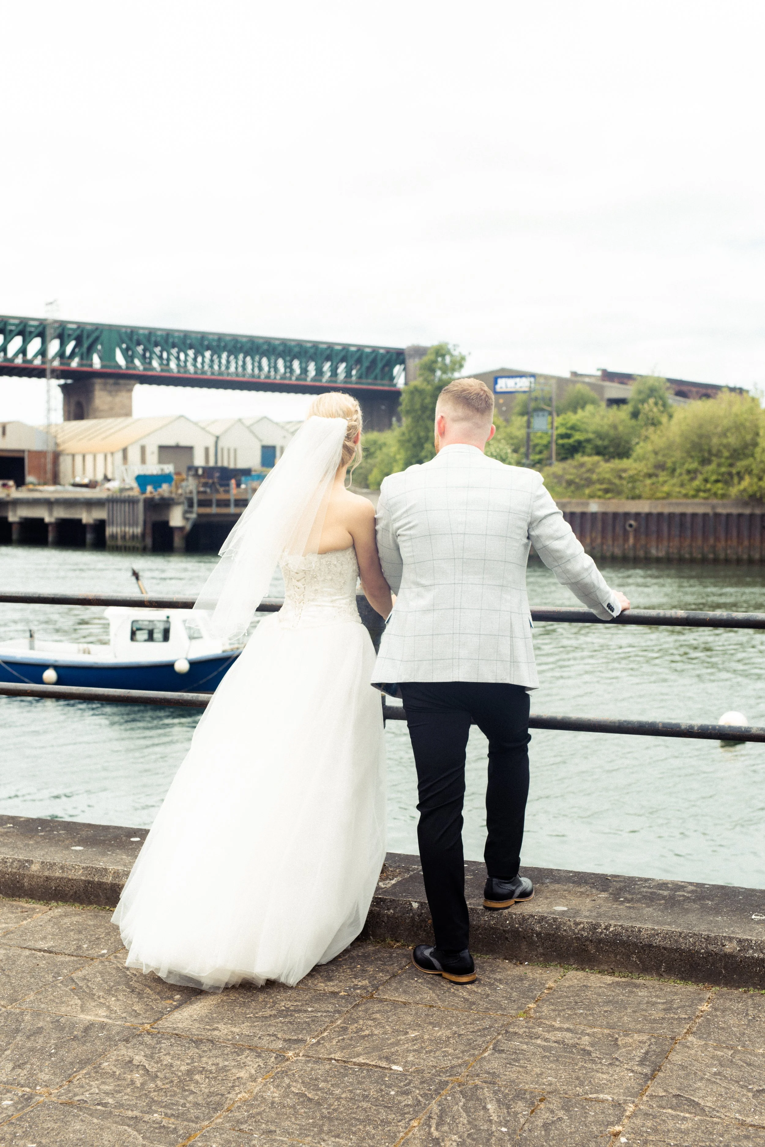 A bride and groom stand by the water's edge with their backs to the camera, looking at the water and a boat nearby.