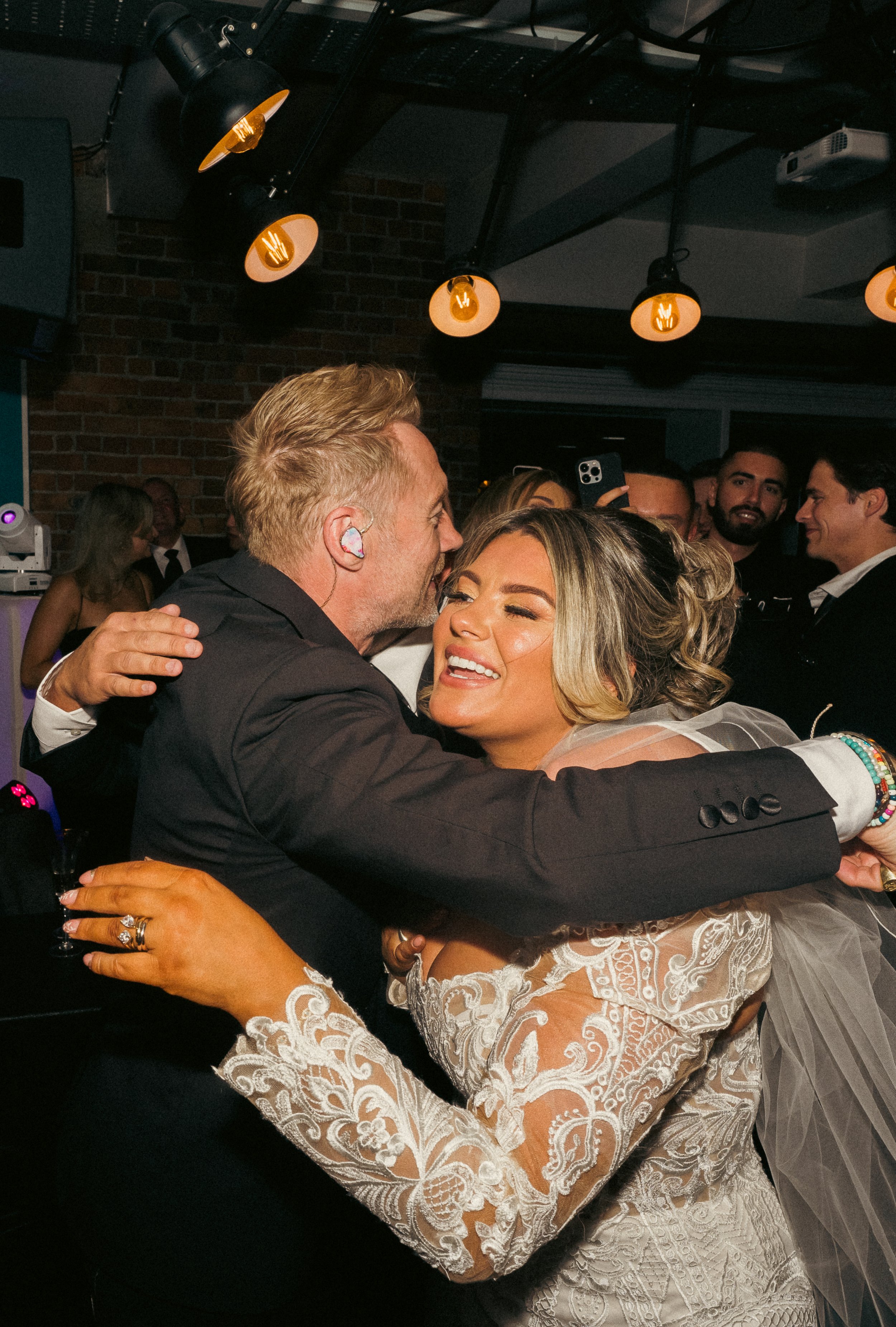 Two people hugging at a celebration, with a woman in a detailed lace dress and a man in a dark suit, surrounded by other guests in a dimly lit venue.
