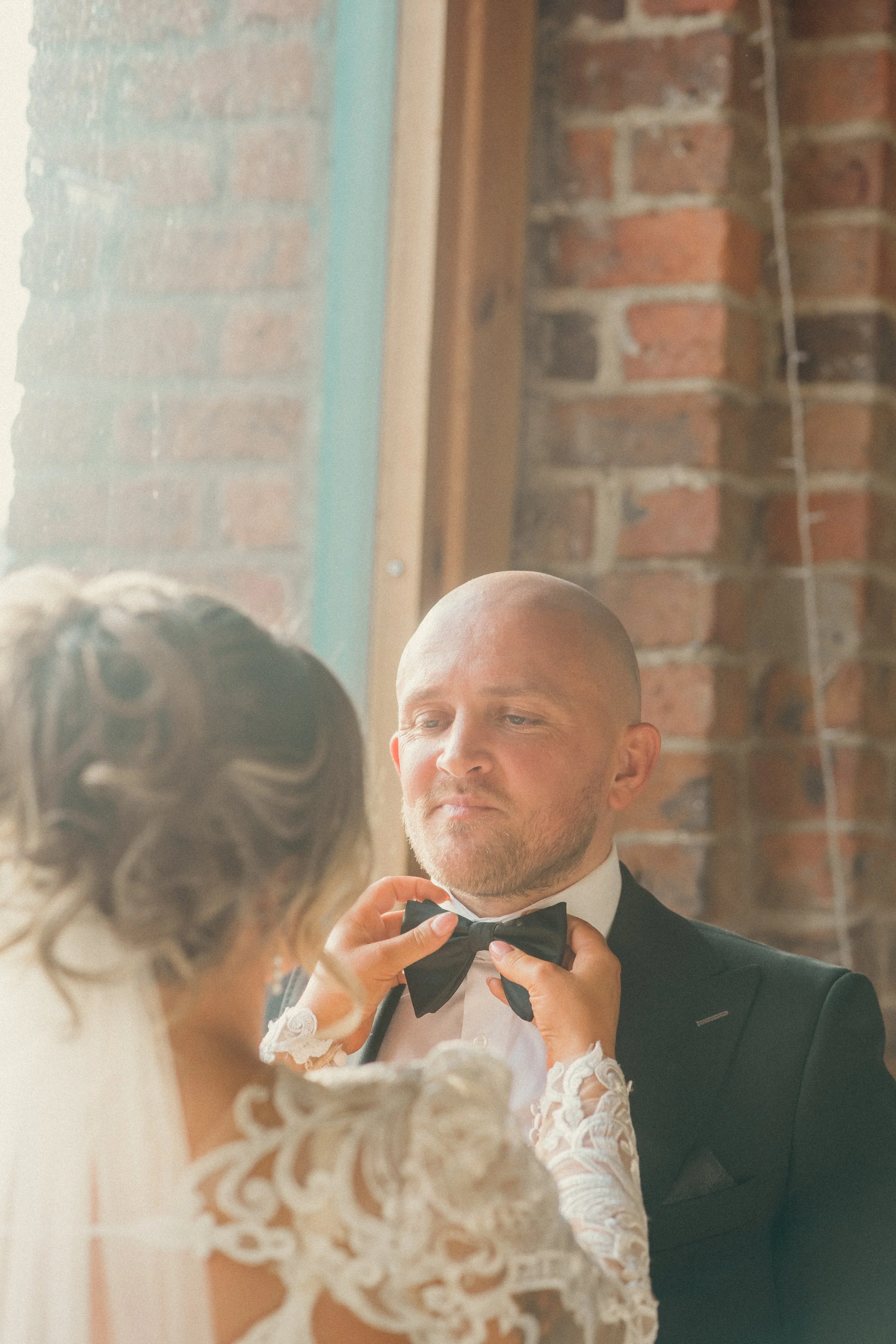 A woman helping a man adjust his black bow tie in a room with brick walls and wooden beams.