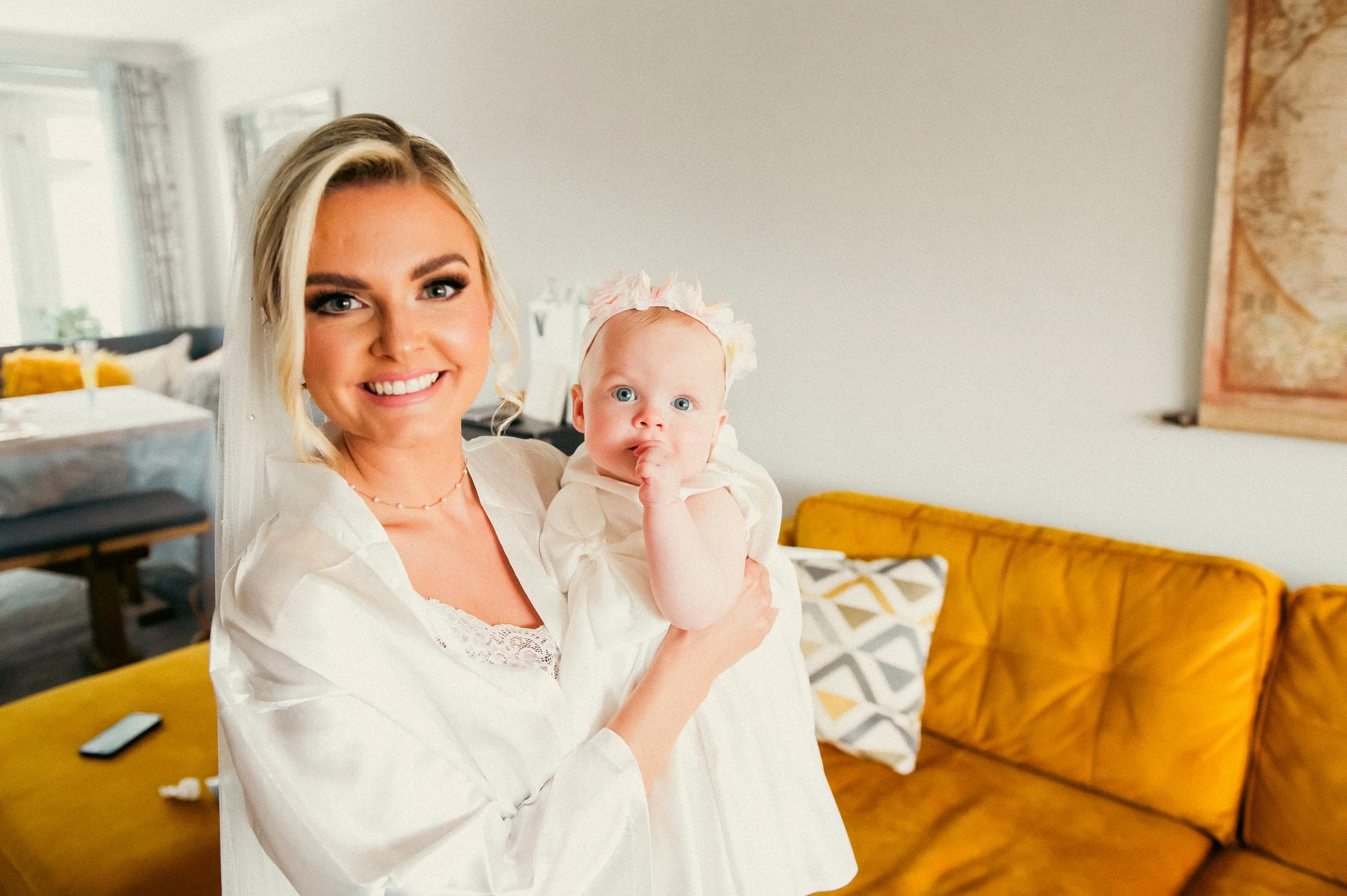 A smiling woman with blonde hair holding a baby girl with blonde hair and blue eyes, wearing a white dress and floral headband, in a living room with yellow sofa and patterned decor.