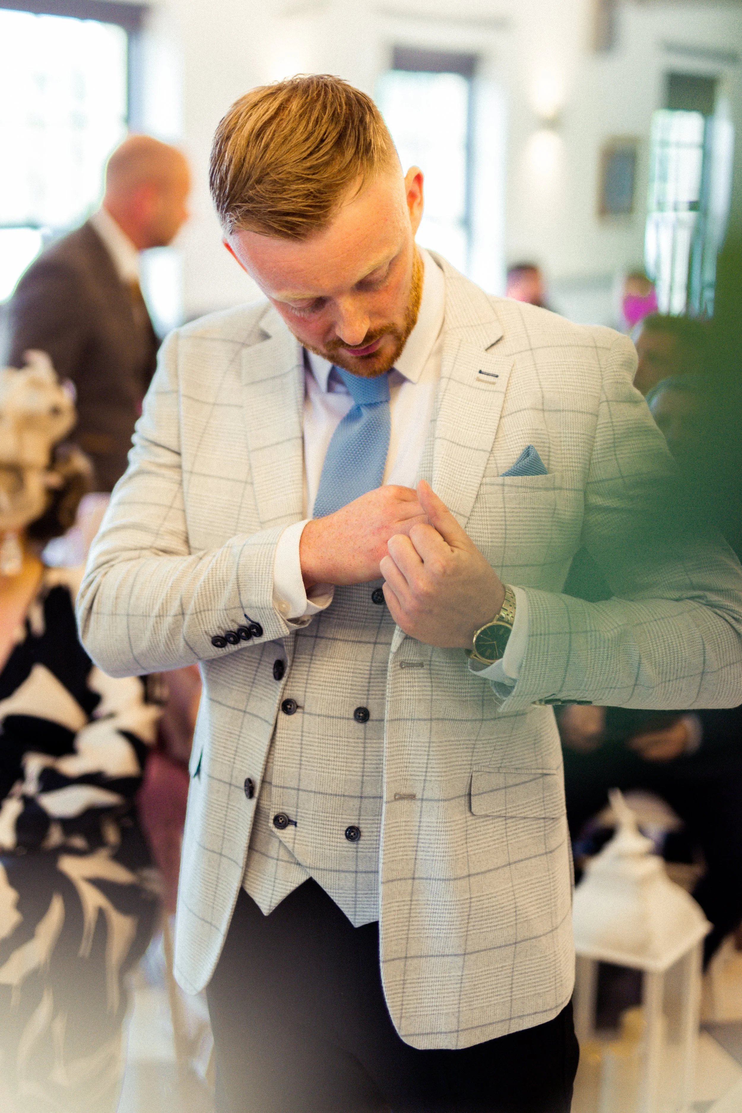 A man in a light-colored plaid suit, blue tie, and watch adjusting his cufflinks in a well-lit indoor setting, possibly at a formal event.