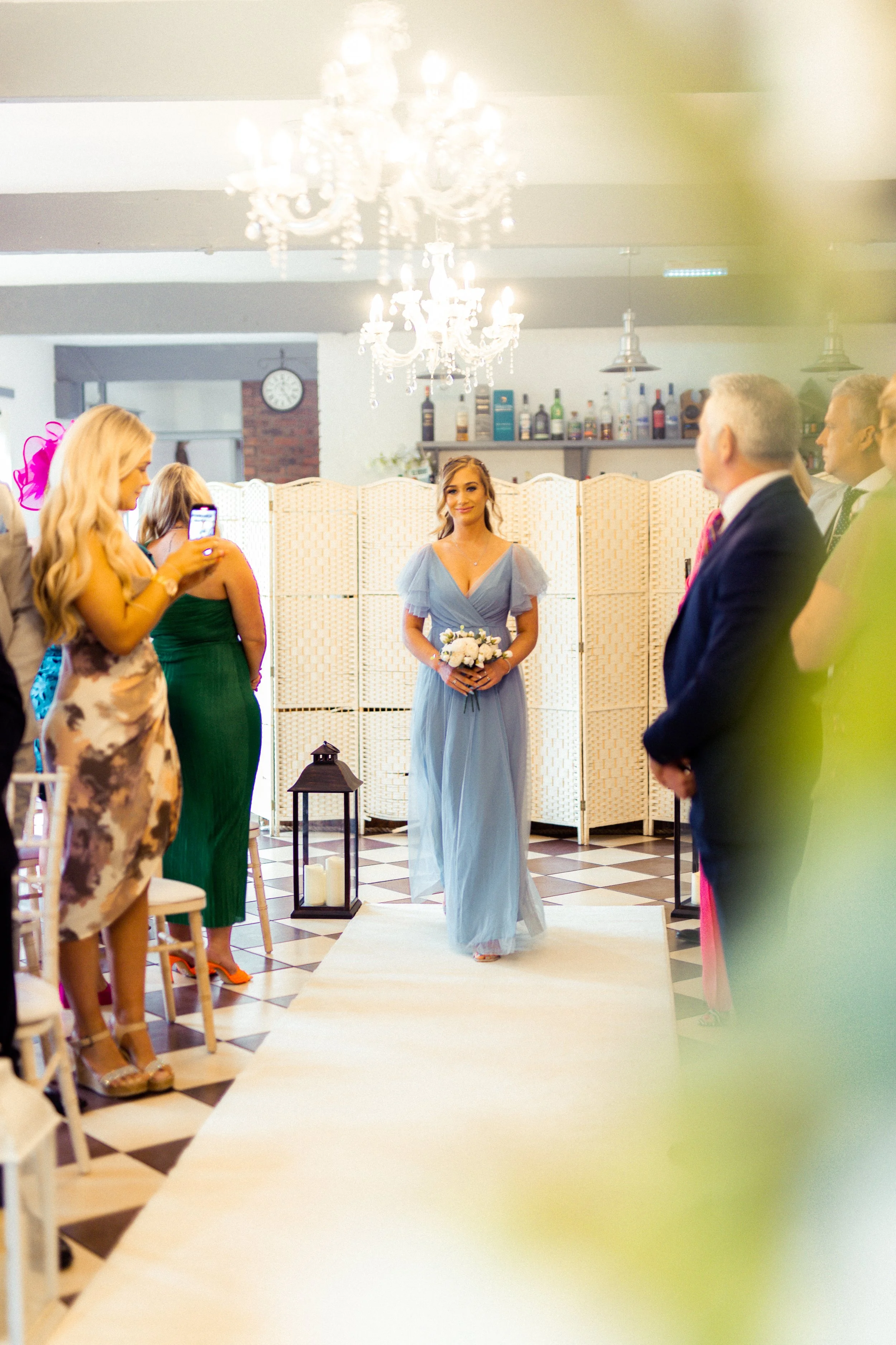 A woman in a long, light blue dress stands holding a bouquet of white flowers, as guests in formal attire watch her during a wedding ceremony in an elegant indoor venue with chandeliers and checkered floor.