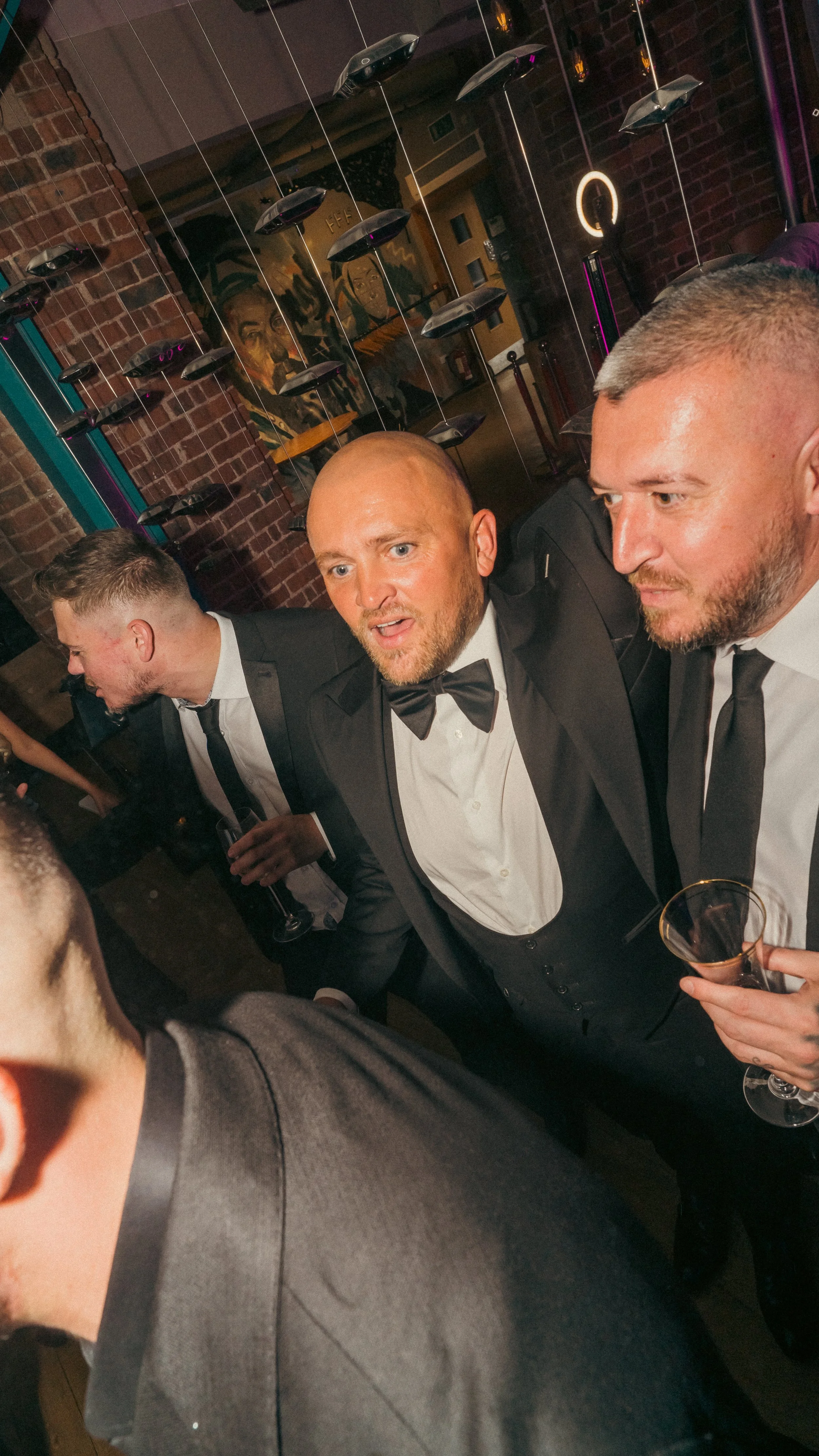Men in tuxedos at a formal party, talking and holding drinks in a dimly lit venue with brick walls and decorative art.