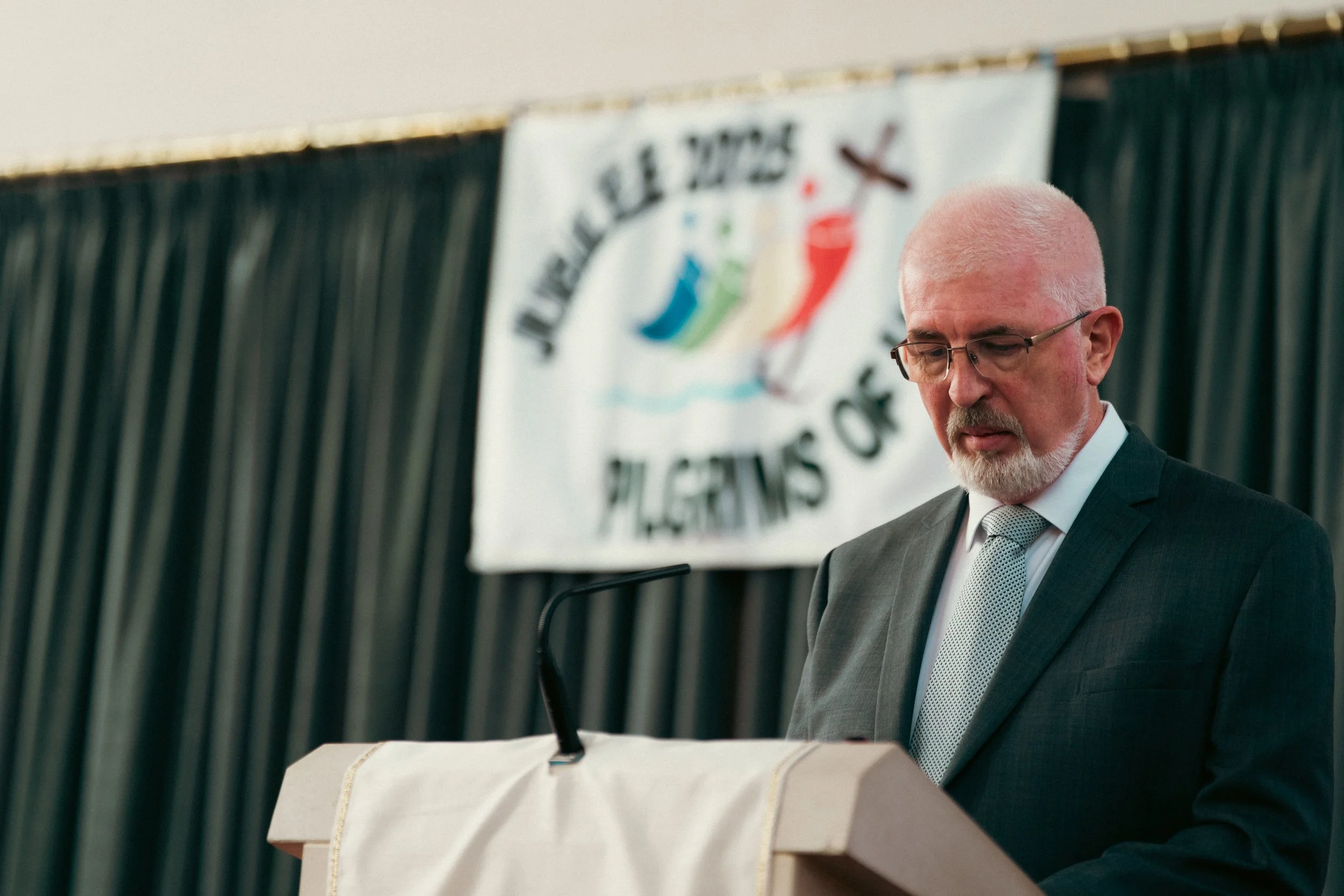 A man with gray hair and glasses wearing a dark suit and tie standing at a podium with a white cloth, looking down. Behind him is a banner with a logo involving colorful figures and a boat.