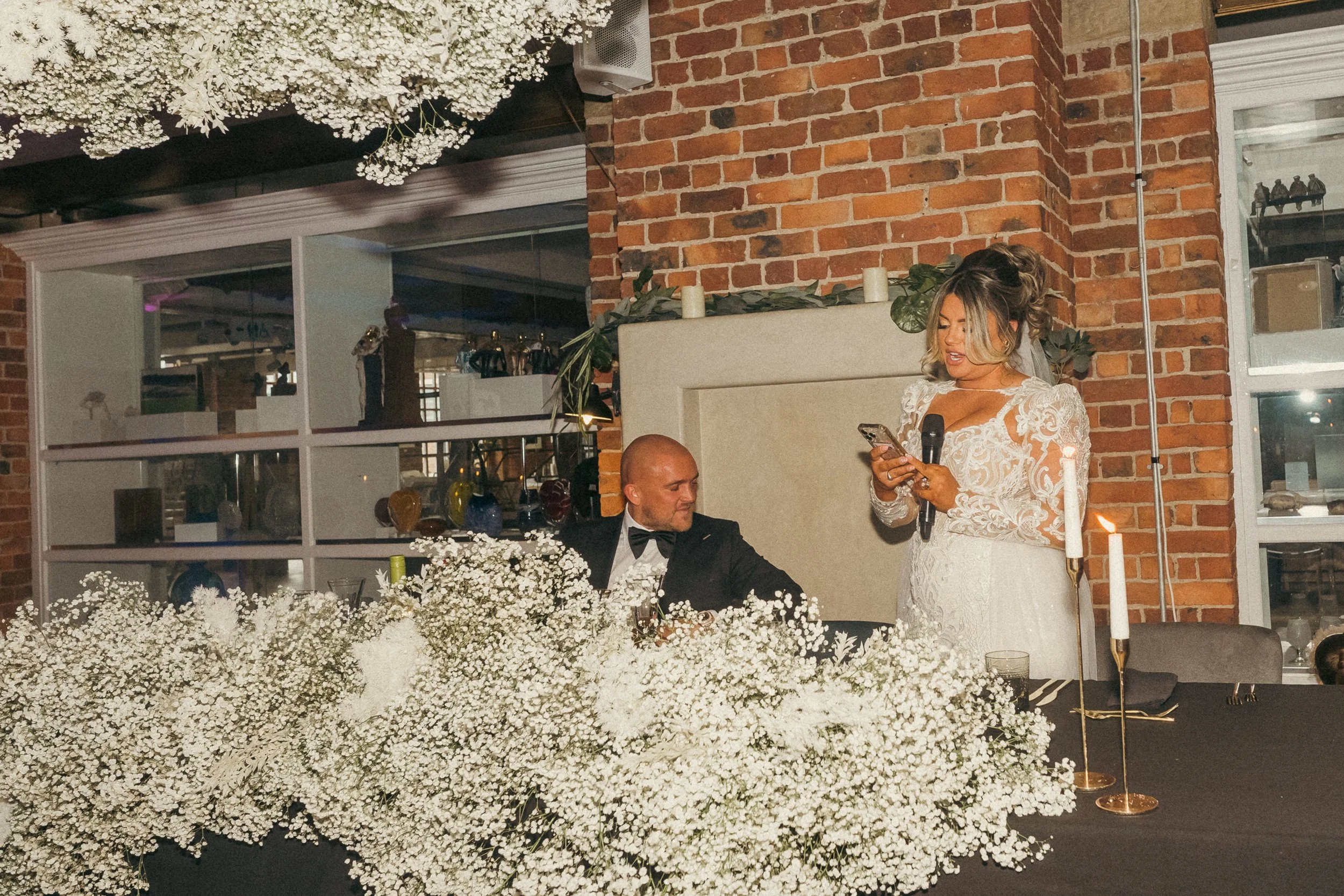 A woman in a white wedding dress giving a speech at a wedding reception, holding a microphone and reading from her phone, while a man in a tuxedo listens.