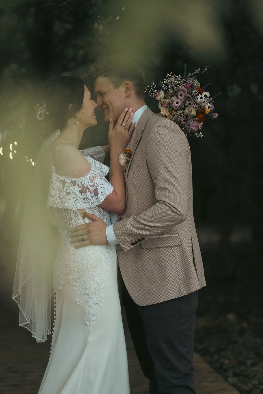 A bride and groom in wedding attire close to each other, smiling and touching faces, outdoors with a blurred background. The bride wears a white lace wedding dress with short sleeves and a veil, while the groom wears a tan suit jacket with a boutonni