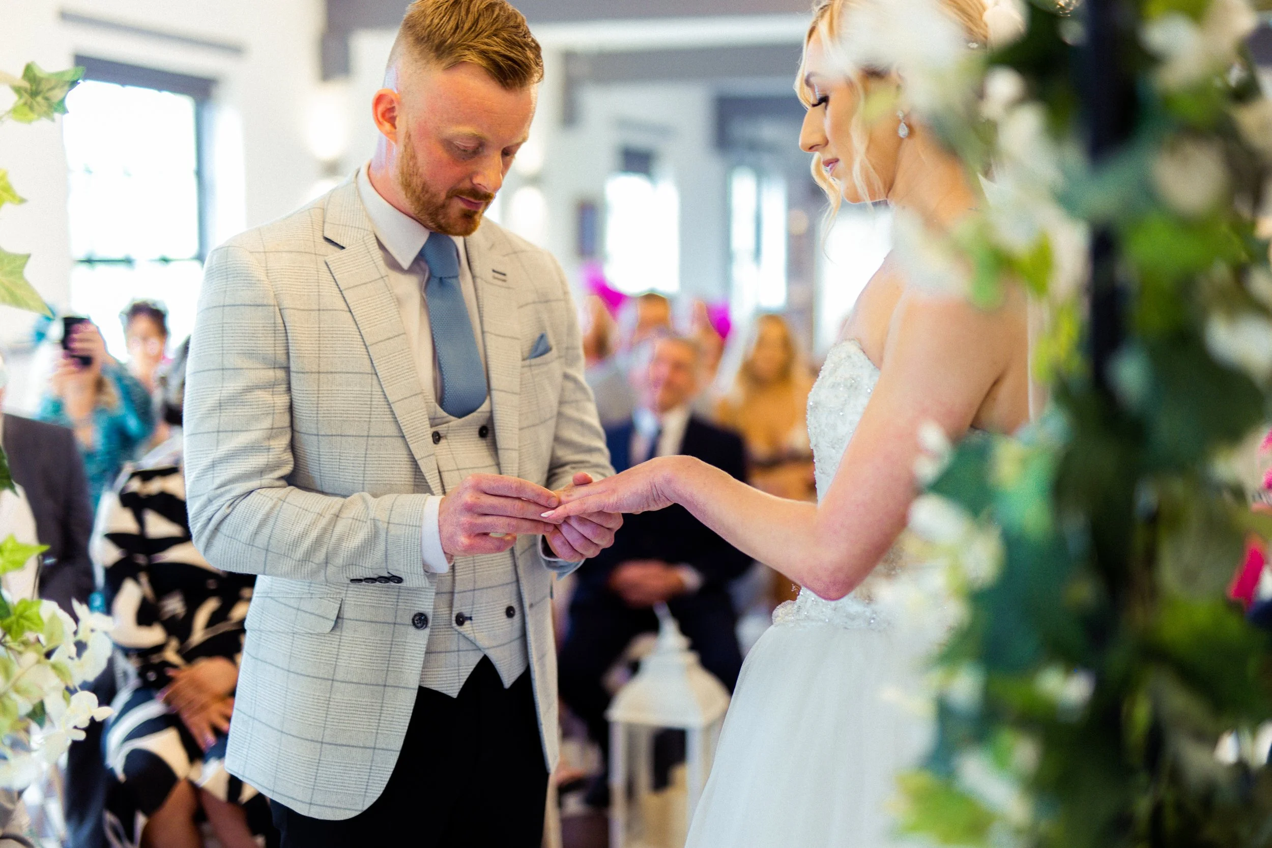 A groom in a light plaid suit and a bride in a white wedding dress exchanging rings during their wedding ceremony, surrounded by guests in a decorated indoor setting.