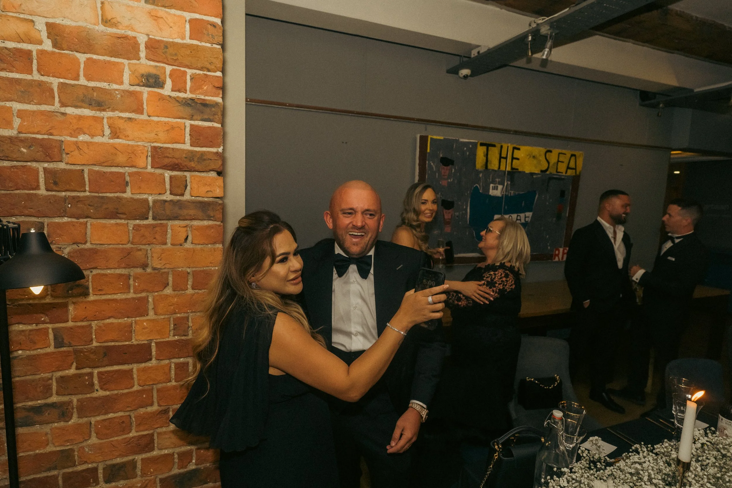 People dressed in formal attire at a dinner party, with a brick wall on the left and a gray wall with a 'The Sea' painting in the background. Some guests are socializing in the background.