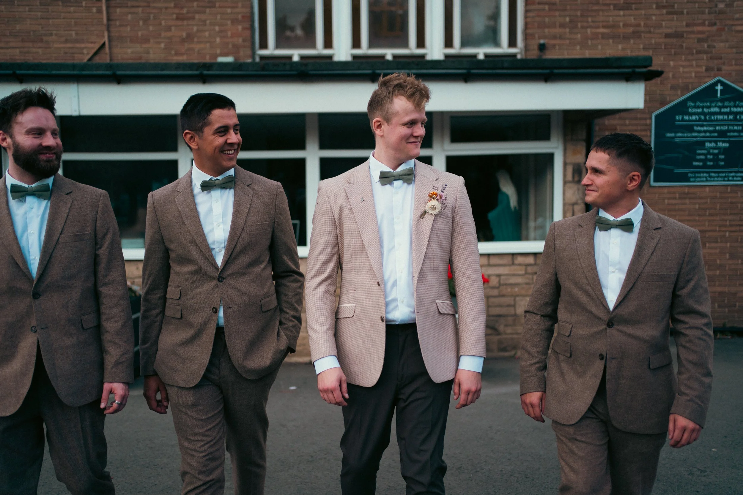 Group of five men dressed in suits and bow ties standing outside a building, smiling and talking.