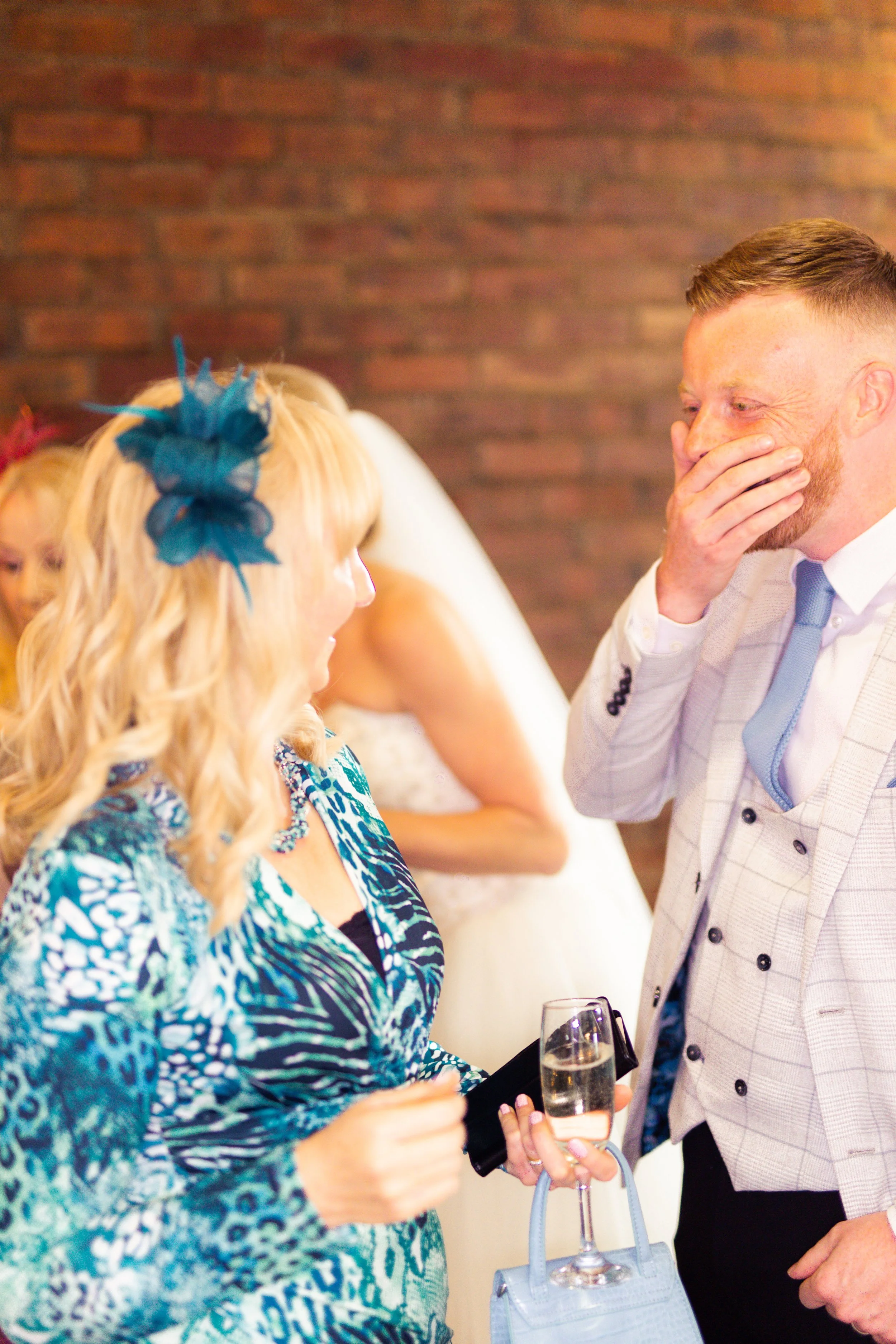 A woman with long blonde hair wearing a blue patterned dress and fascinator converses with a man in a light-colored checkered suit, holding a glass of champagne at a social event.