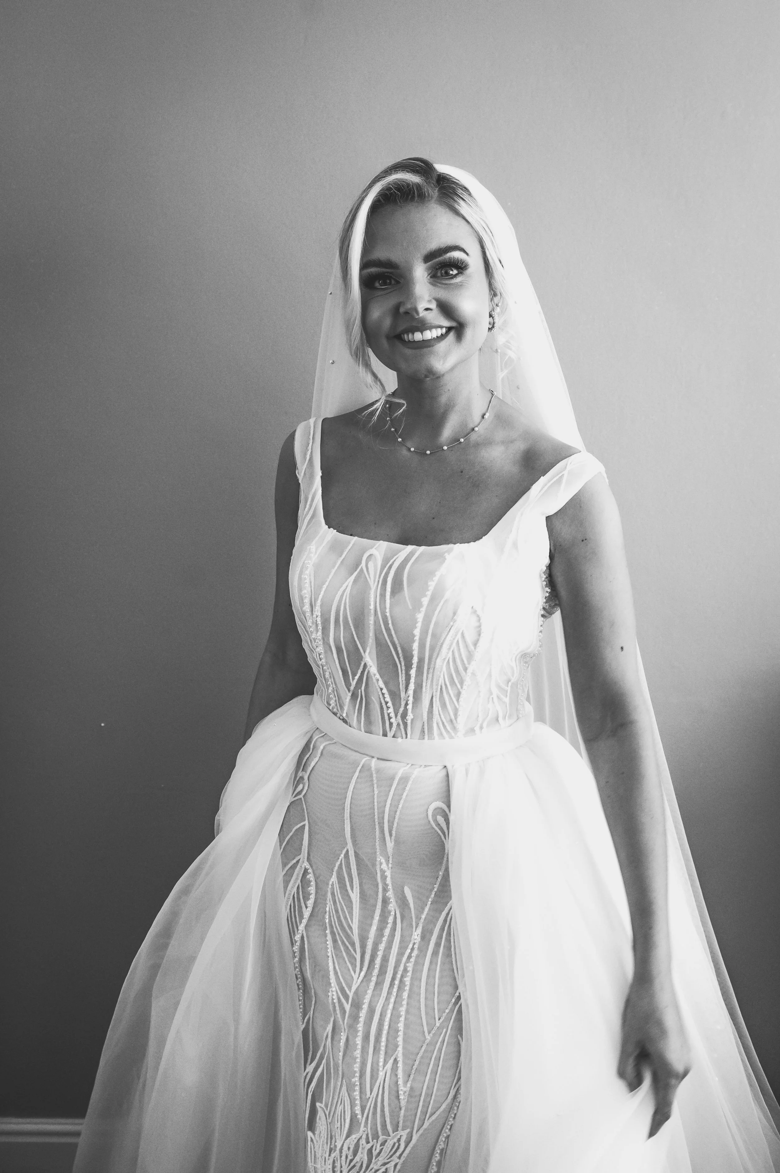 Black and white photo of a smiling woman in a wedding dress with a veil, standing against a plain wall.