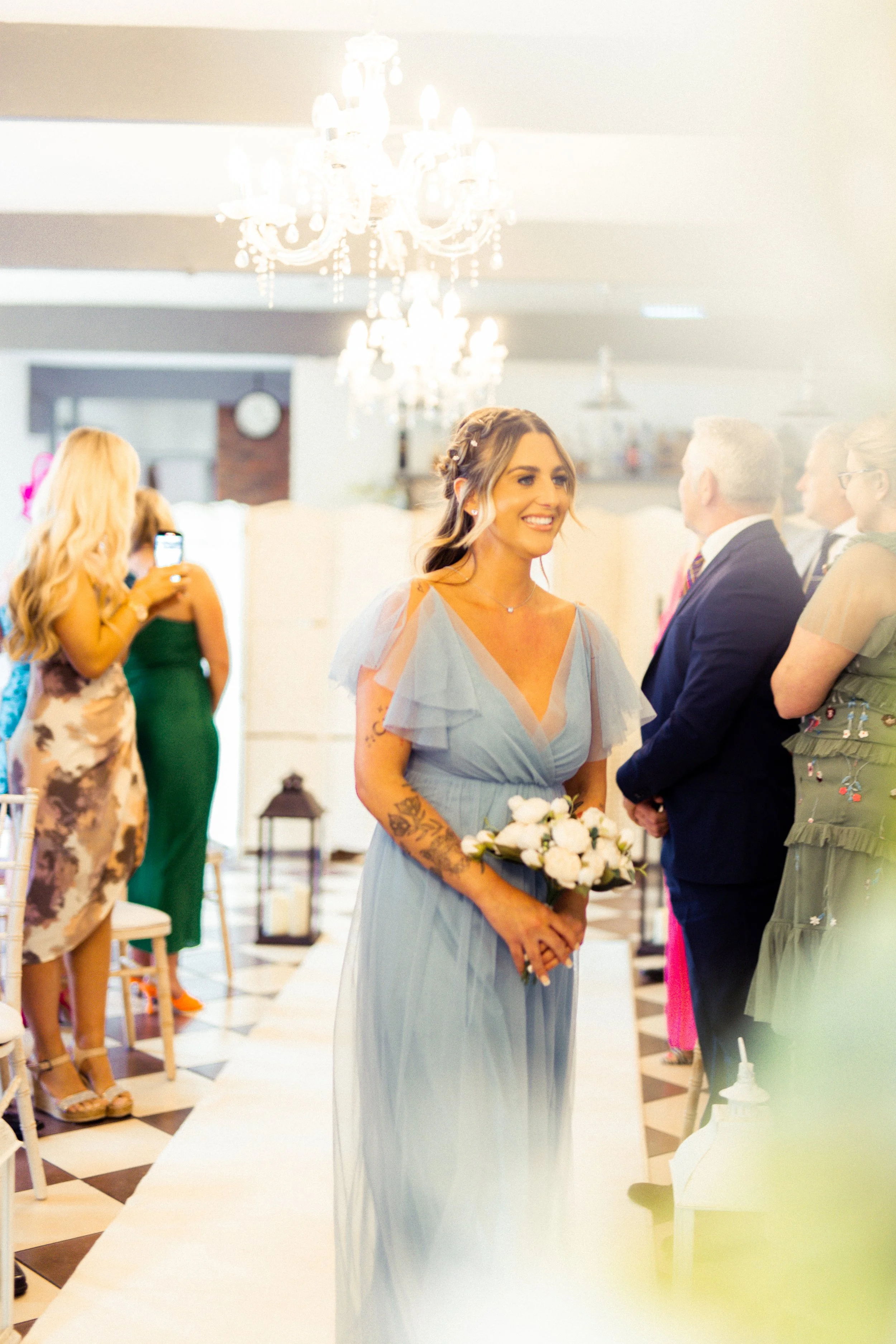 A woman in a light blue dress holding a bouquet of white flowers at a wedding ceremony, smiling amidst guests in formal attire.