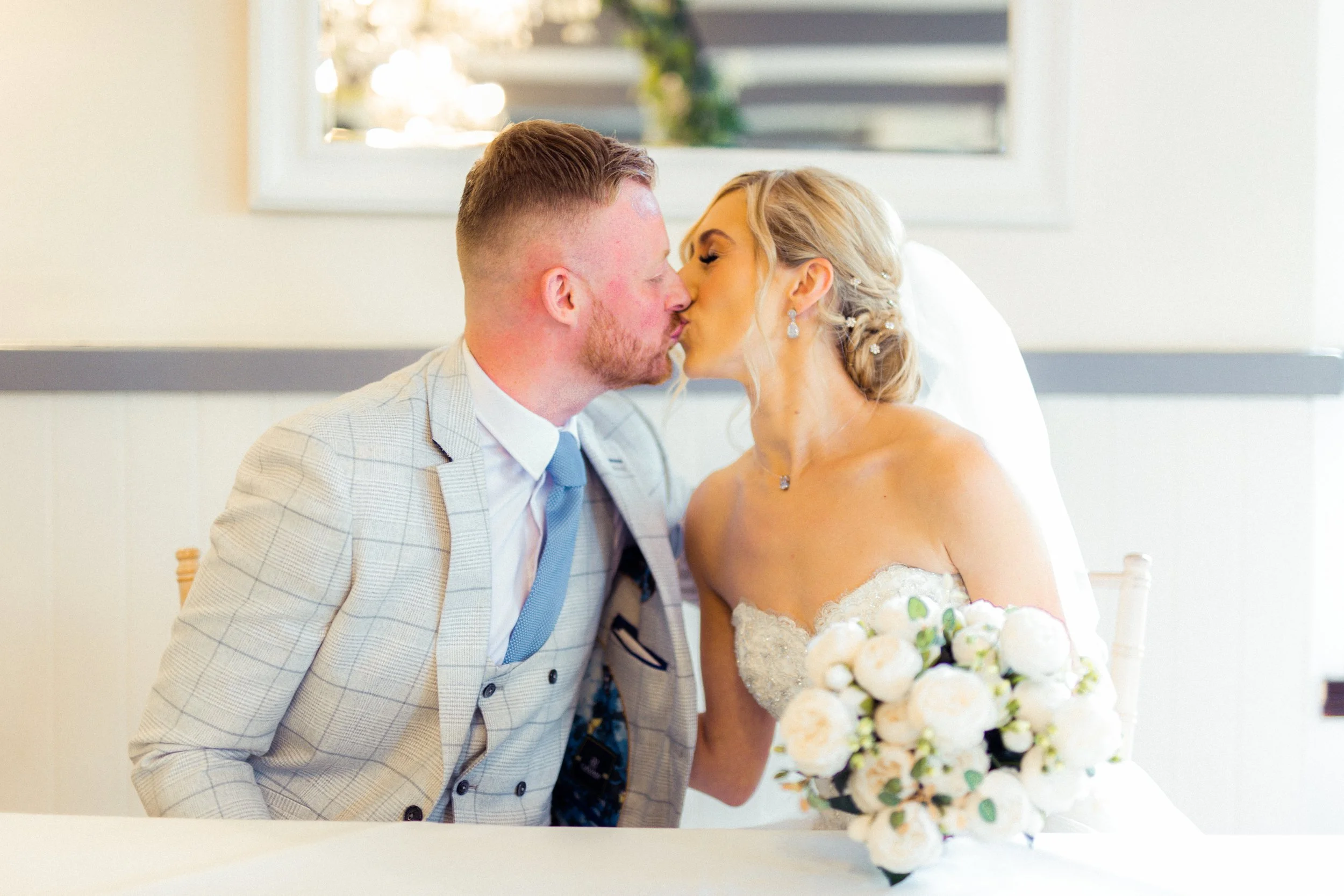 A bride and groom sharing a kiss at their wedding, with the bride holding a bouquet of white flowers, indoor setting with bright lighting and a window in the background.