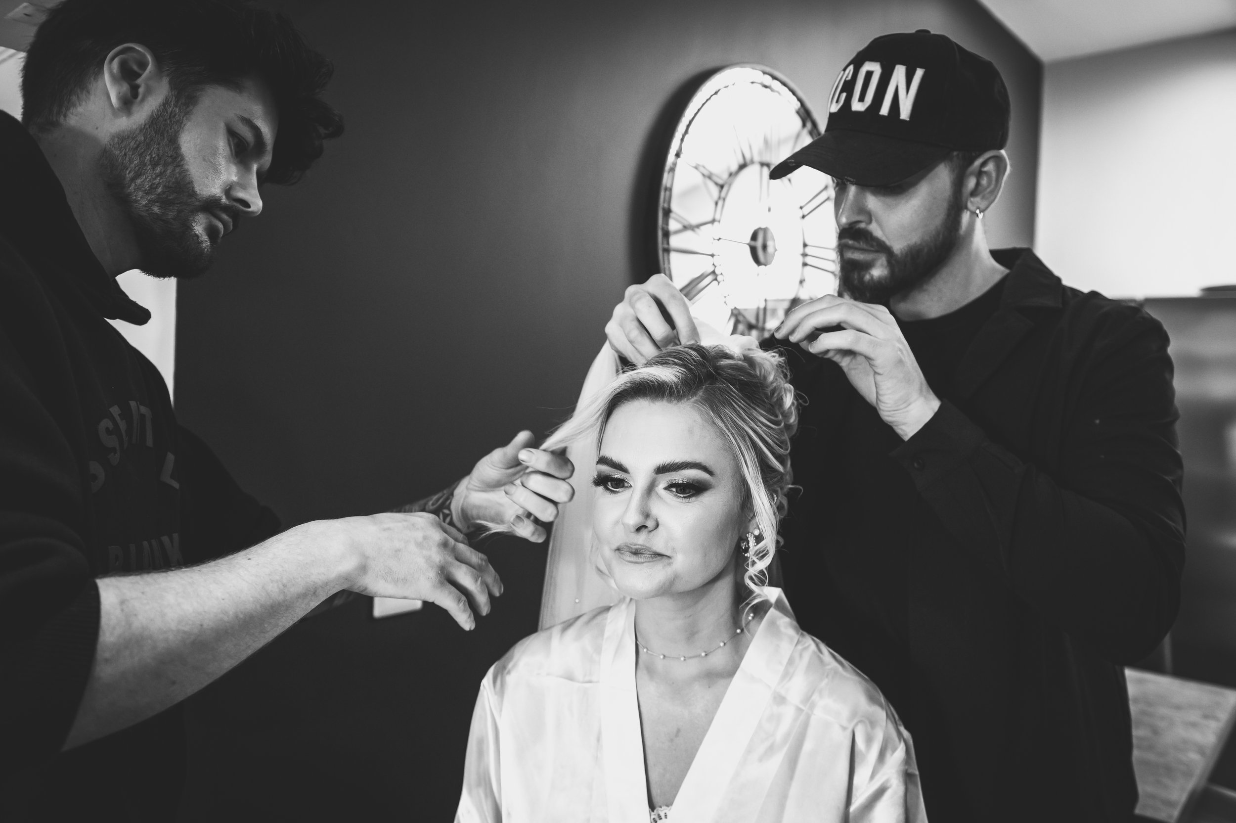 A woman with styled blonde hair sitting as two male hairstylists work on her hair inside a salon. One stylist is on her left, holding a strand of her hair, and the other on her right, adjusting her hairstyle. The woman wears a satin robe, earrings, a