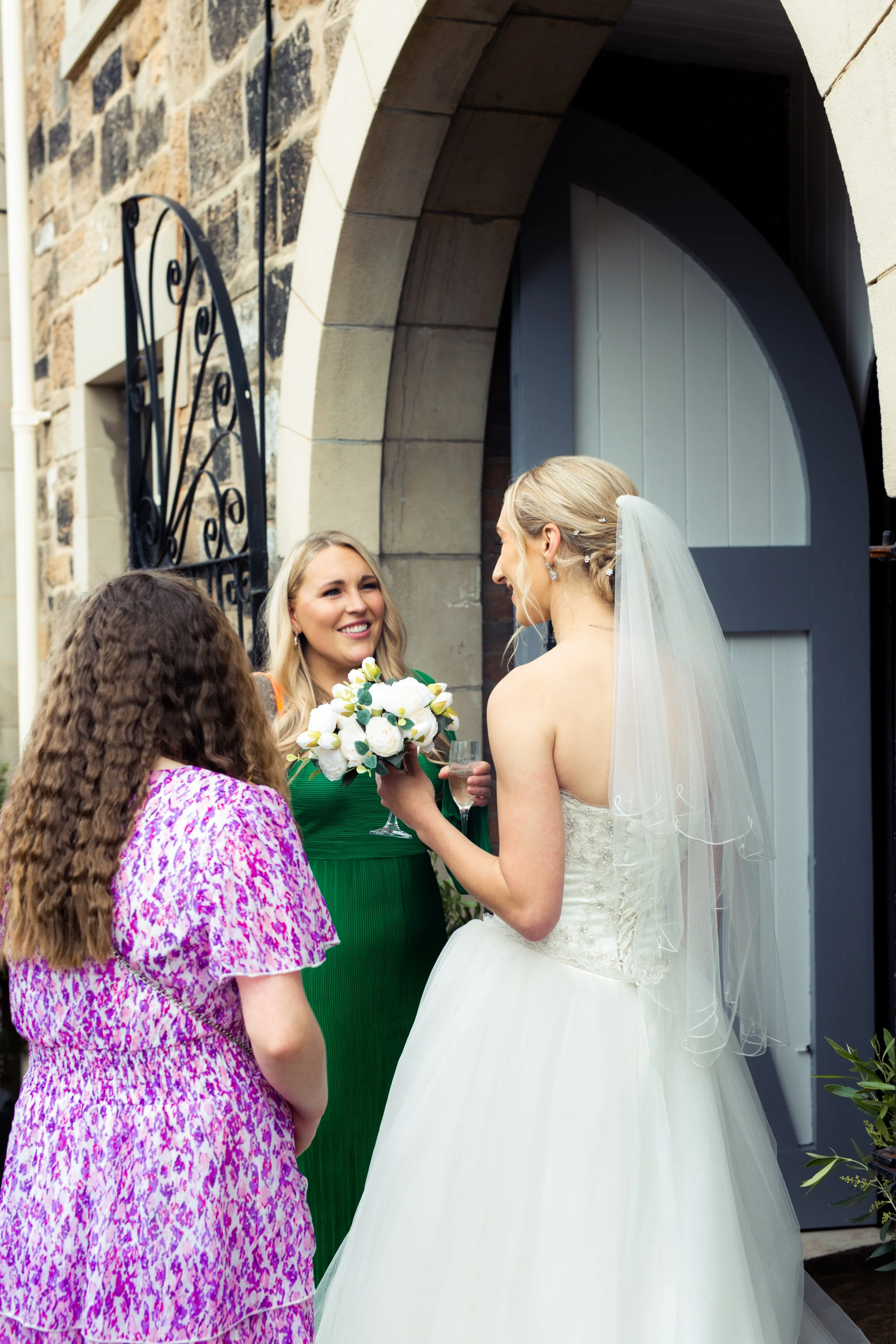 A bride with a veil and strapless wedding dress smiling and holding a bouquet, talking to two women outside a building with a stone archway and blue door. One woman wears a green dress and holds a wine glass, the other a pink patterned dress.