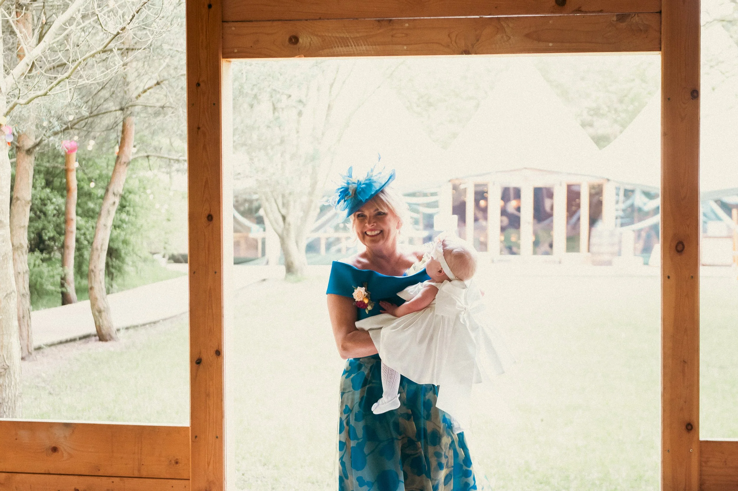 A woman dressed in a blue dress and matching hat holding a baby in a white dress outdoors near a wooden structure with trees and a playground in the background.