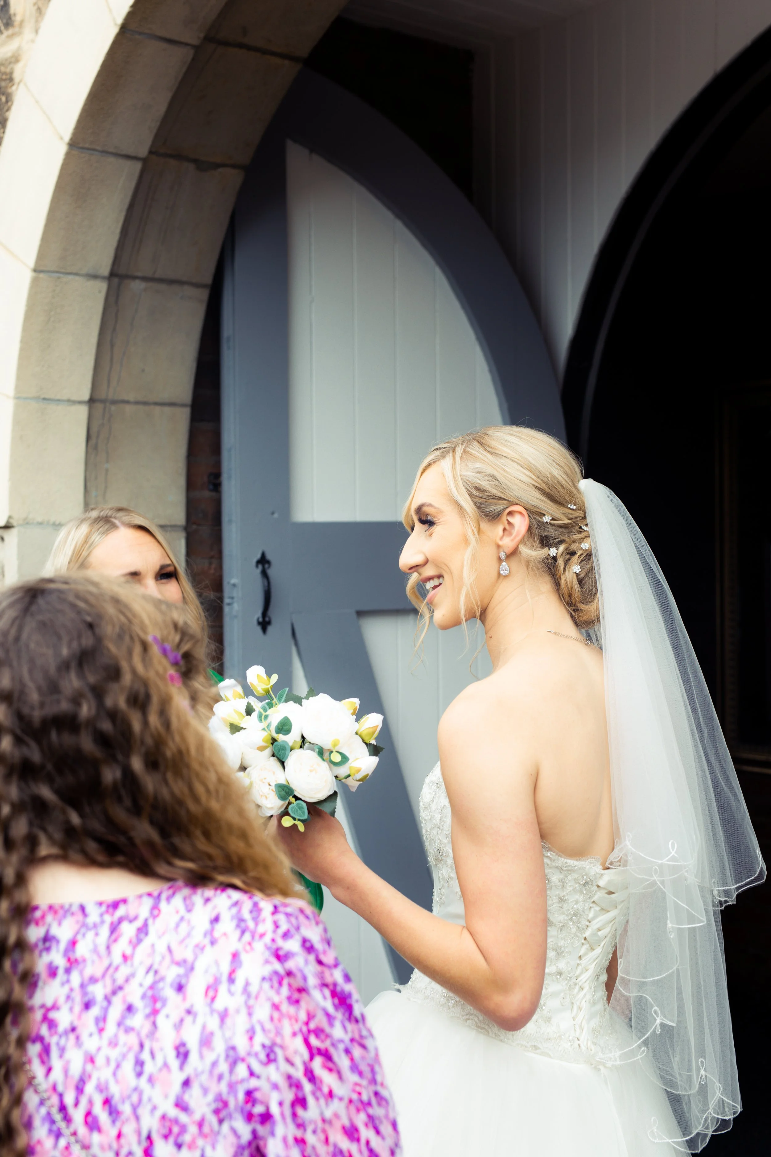 A bride in a white wedding dress and veil smiling and holding a bouquet, interacting with women outside a building with grey and stone archway doors.