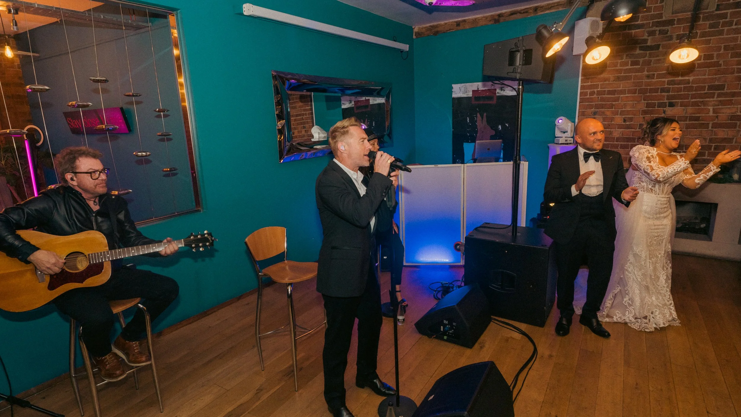 A band performing at a wedding reception with three singers and a guitarist. The singers include a man in a tuxedo and a woman in a lace wedding dress. The guitarist is seated, wearing a black jacket and glasses. The setting is a decorated indoor ven