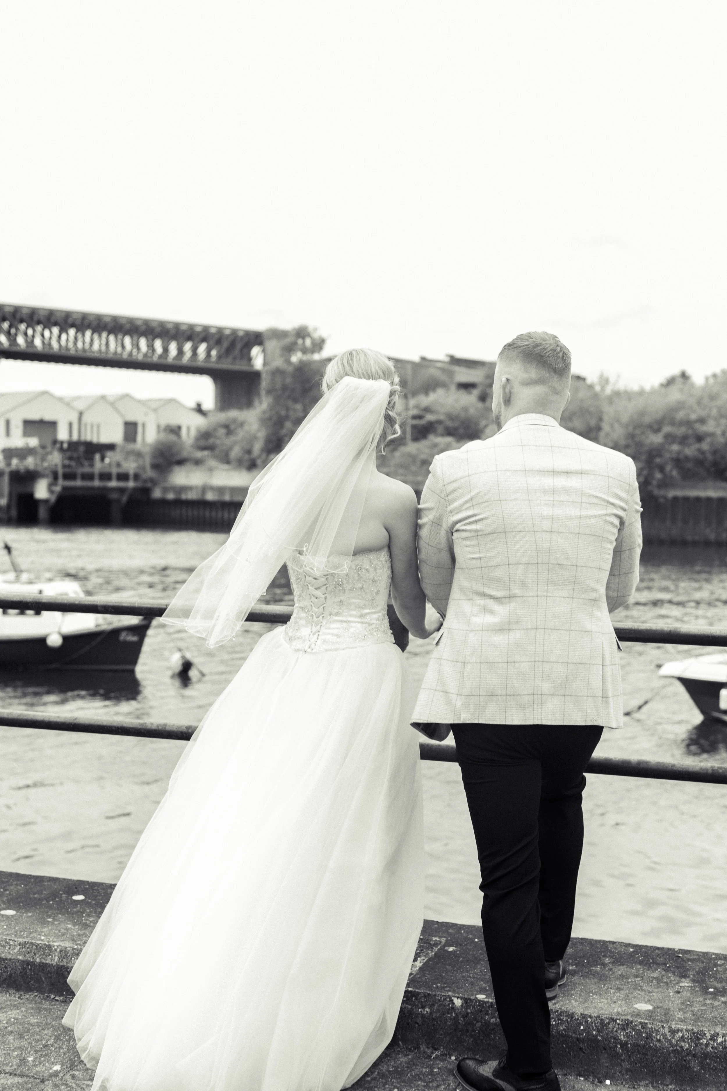 A black-and-white photo of a bride and groom standing by a river, with the bride in a wedding gown and veil, and the groom in a checked suit jacket, looking at the water.
