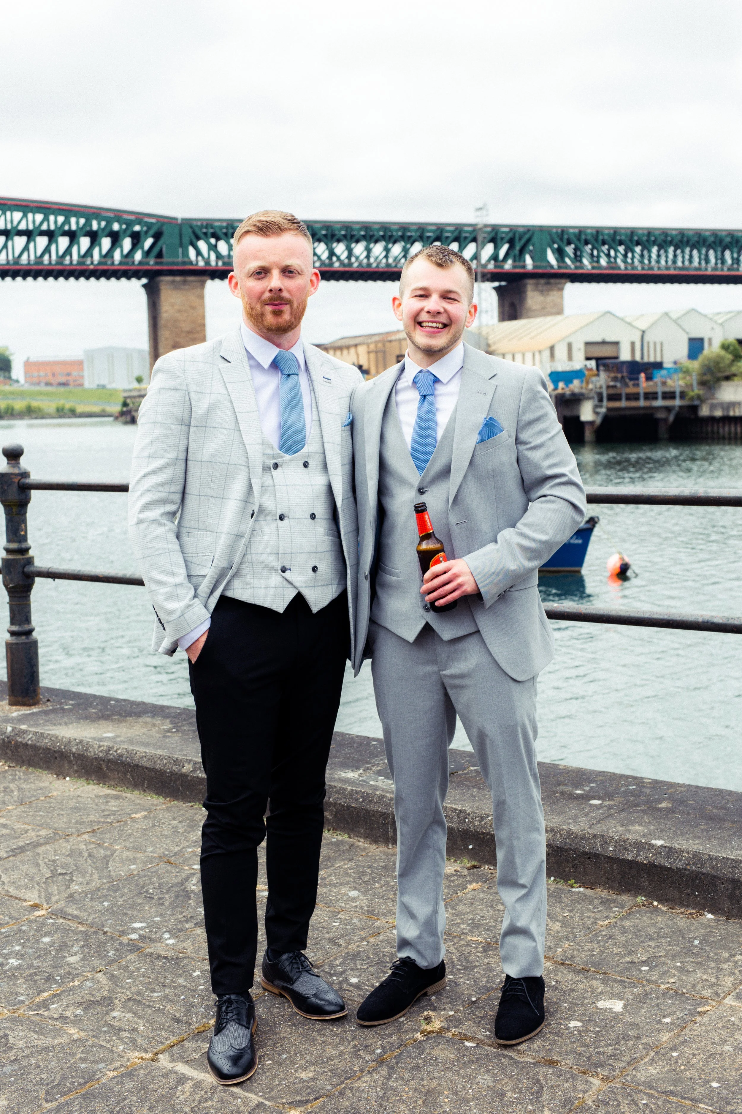 Two men in suits standing by a waterfront with a bridge in the background, smiling; one holding a beer bottle.
