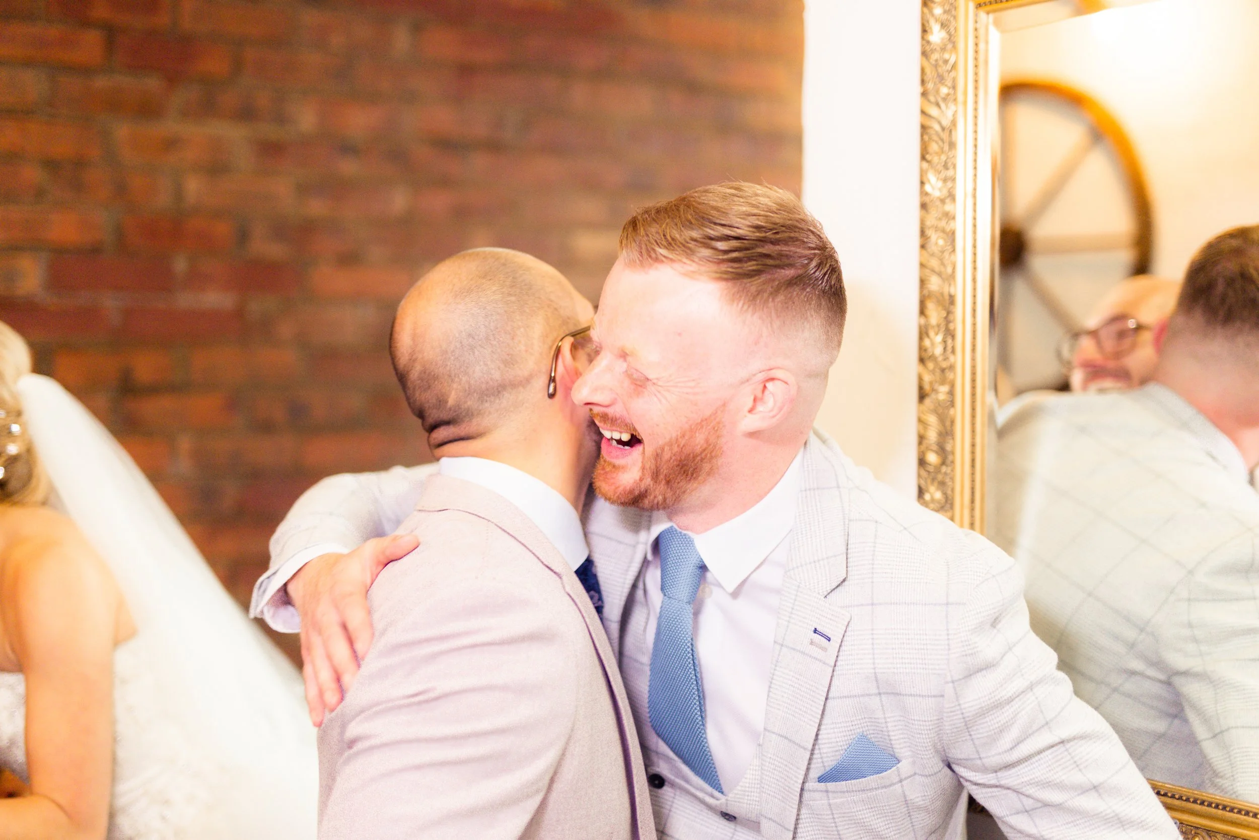 Two men in suits hugging and smiling at a celebration or wedding, with a woman in a white dress and a group of people reflected in a mirror behind them.