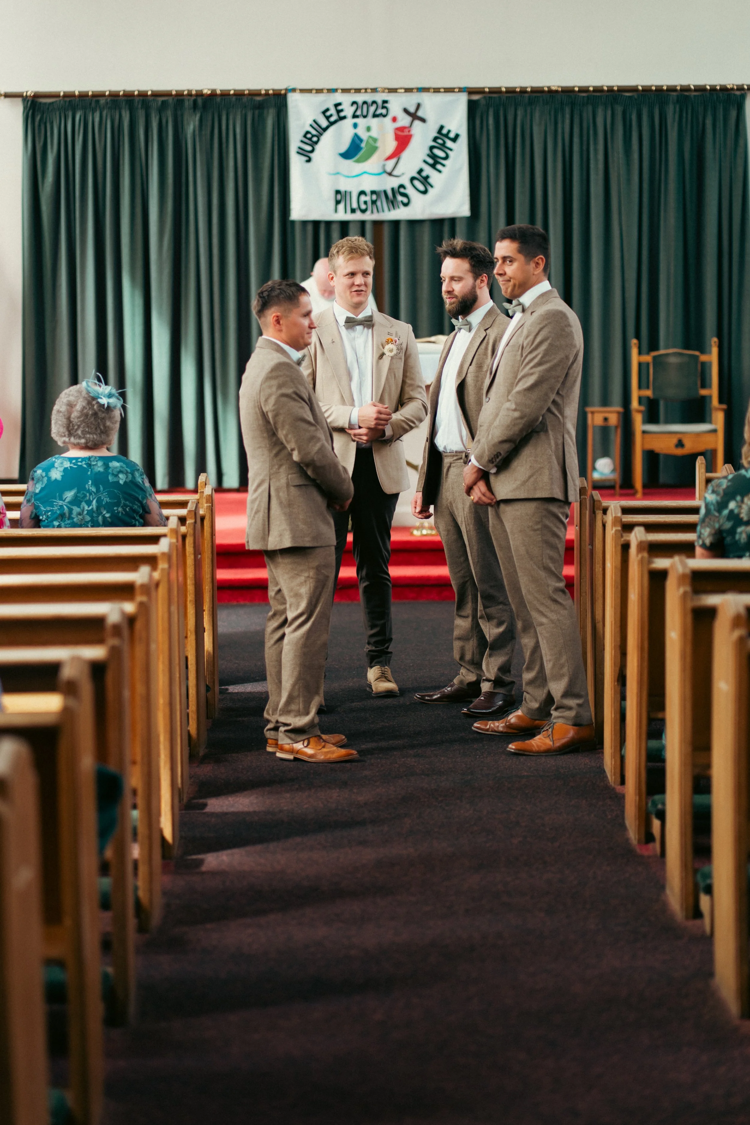 Four men in beige suits standing at the front of a church during a wedding ceremony, with a woman in blue seated to the left and a banner reading 'Jubilee 2025 Pilgrims of Hope' hanging on the green curtain backdrop.