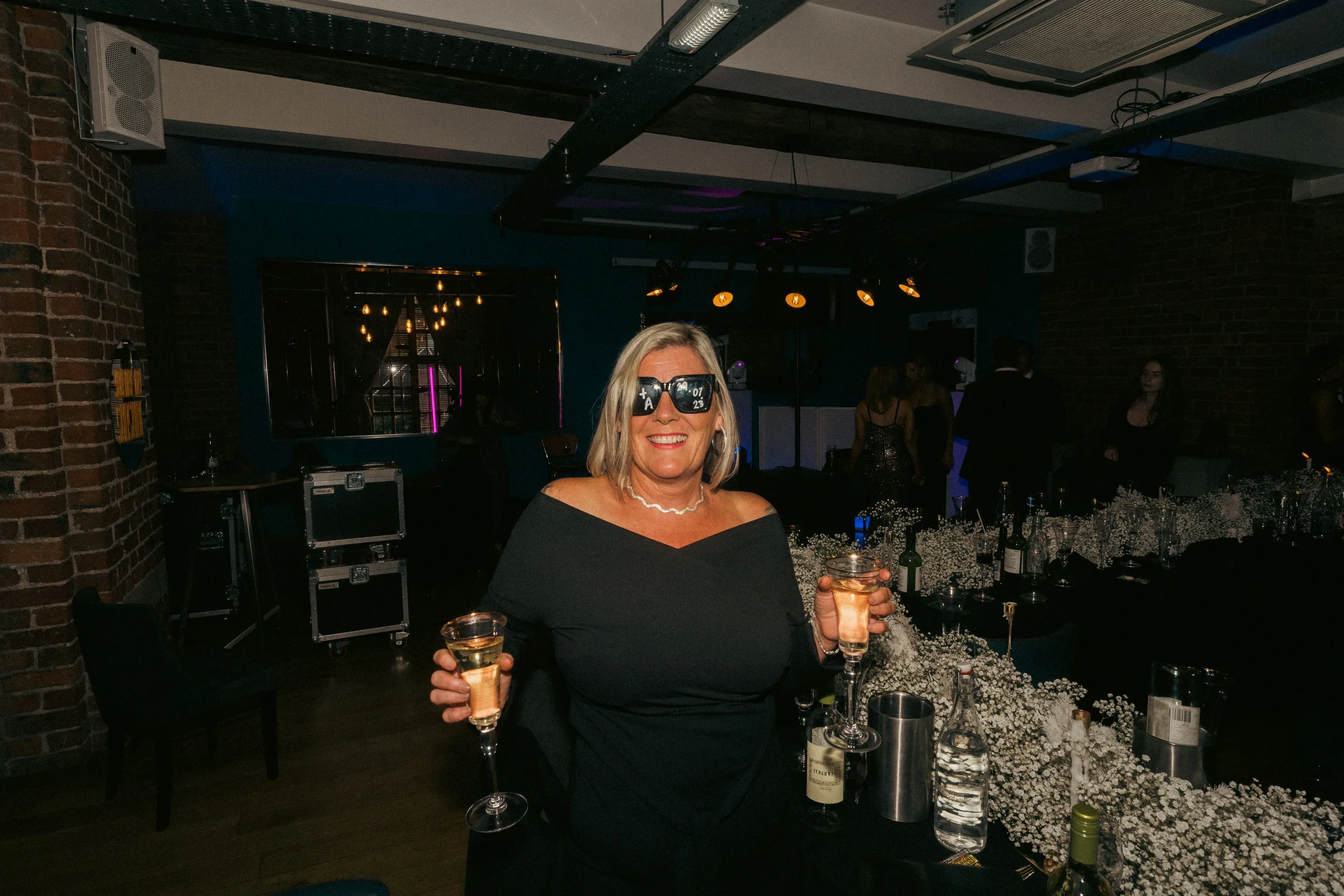 A woman wearing sunglasses and a black off-the-shoulder dress is smiling and holding a glass of champagne at a party or celebration in a dimly lit venue with a brick wall and other guests in the background.