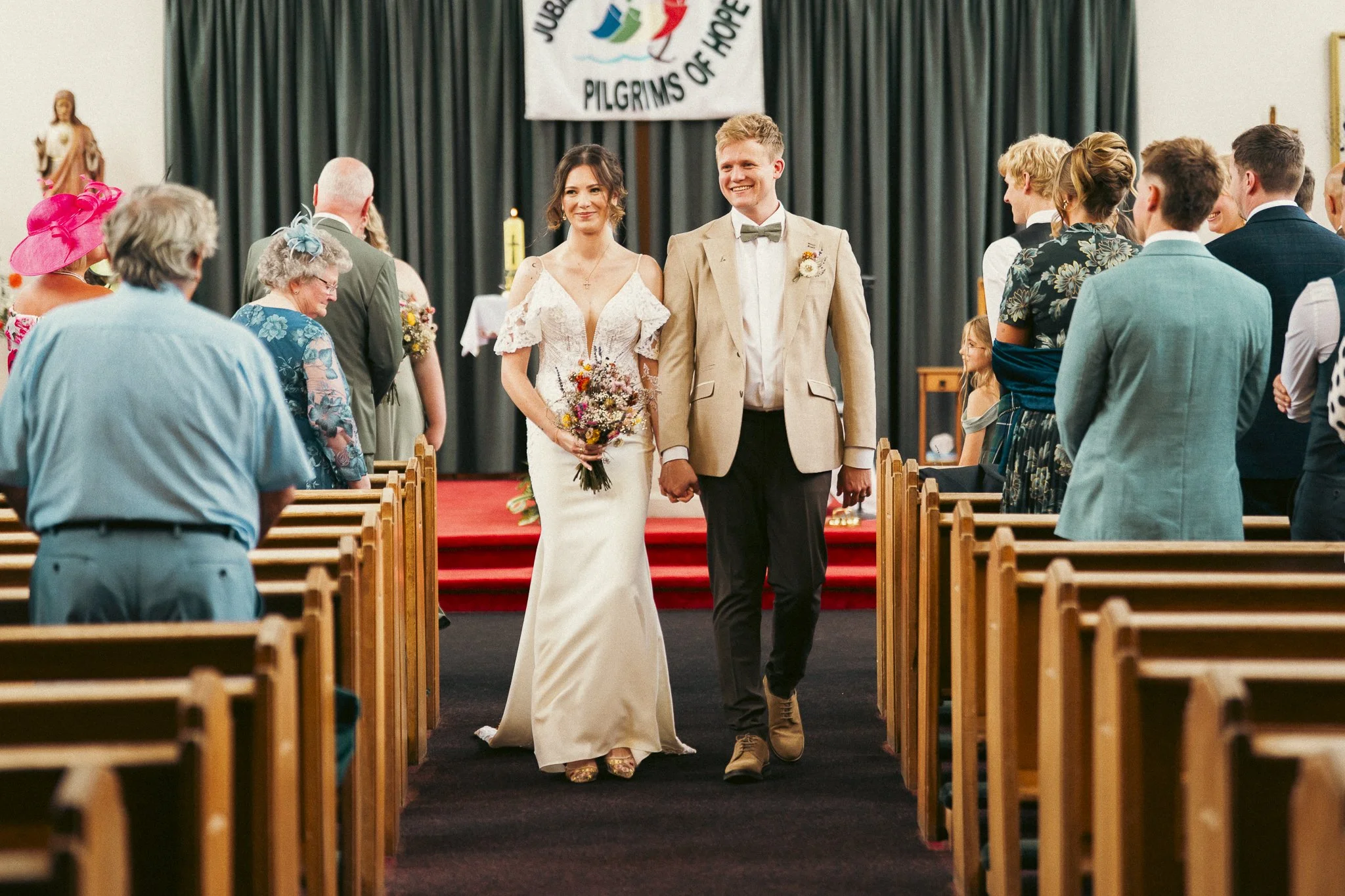 A newlywed couple walking down the aisle after their wedding ceremony, holding hands in a church, with guests standing and clapping.