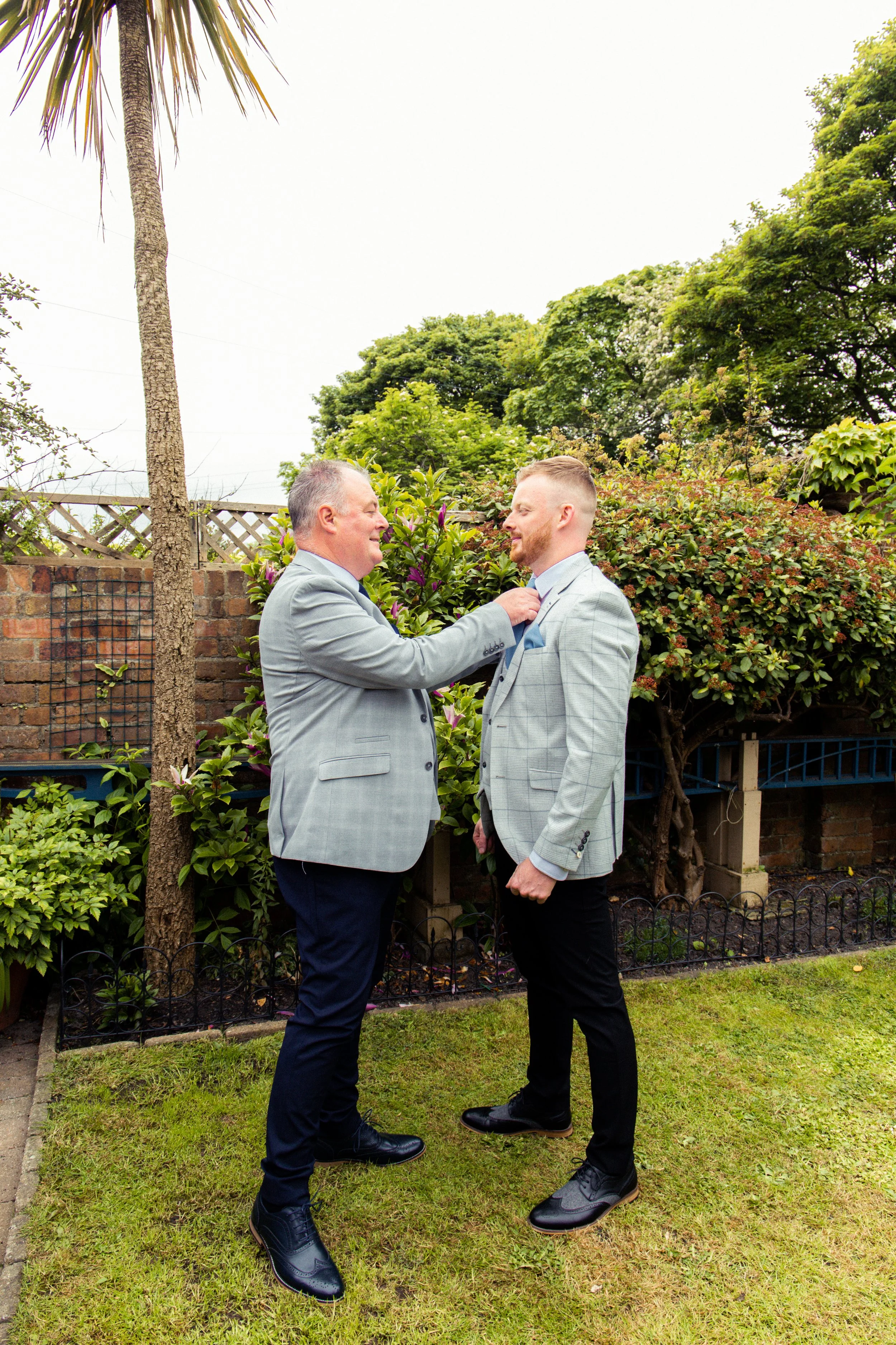 A man helping another man adjust his tie in a garden with trees and bushes.
