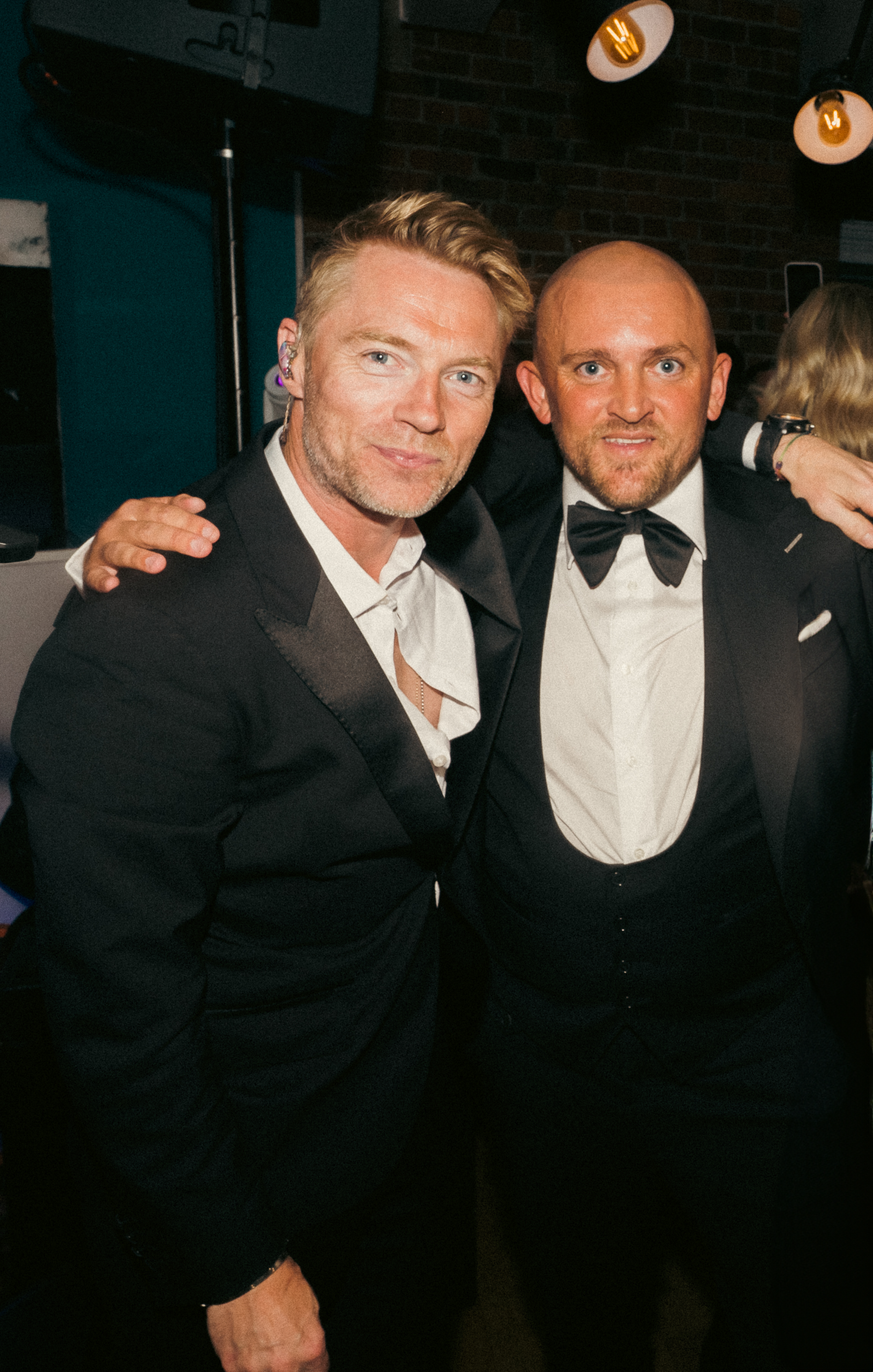 Two men in tuxedos posing together at a formal event, indoors with a brick wall and hanging lights in the background.