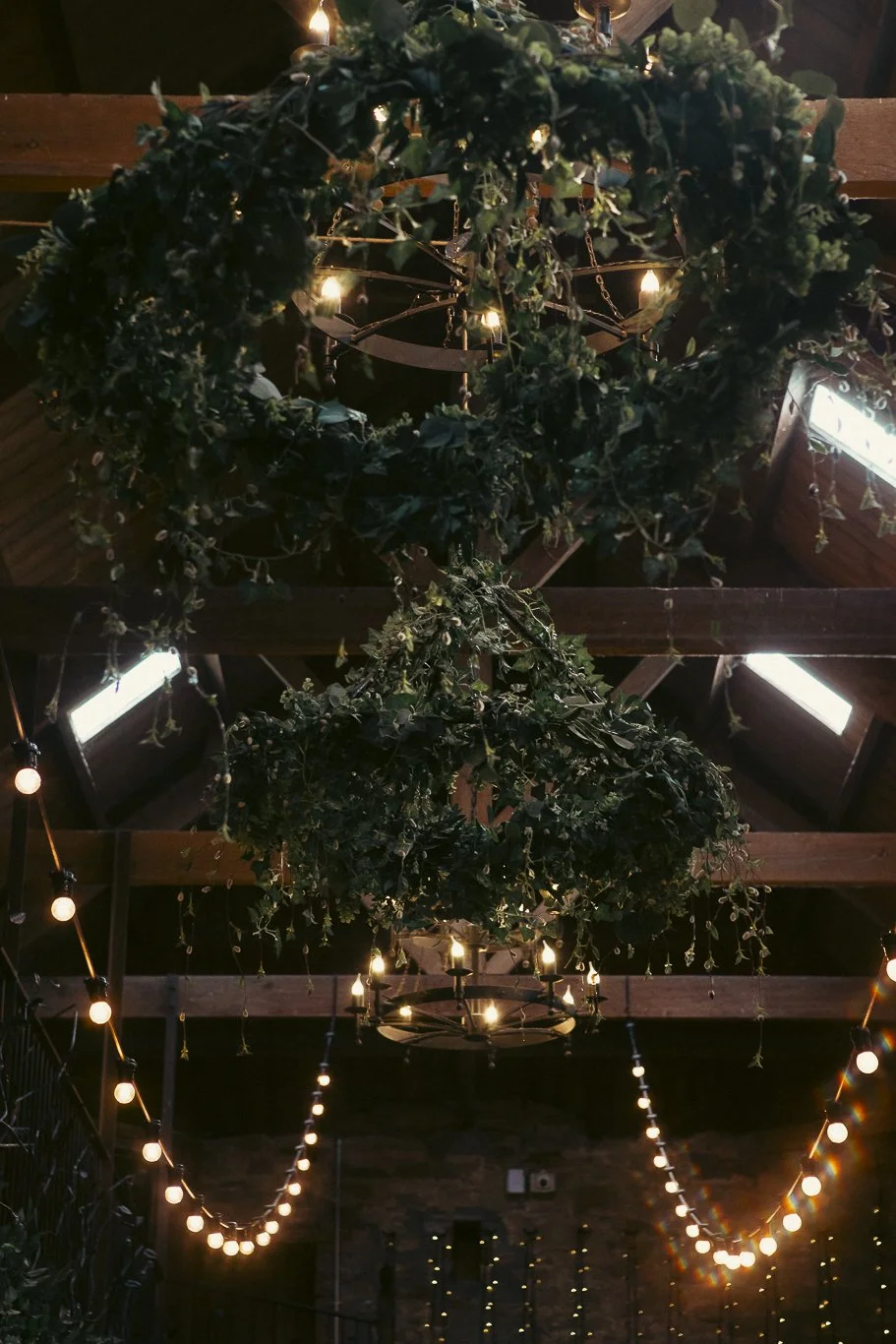 Chandeliers decorated with greenery and hanging light bulbs in a rustic indoor space.