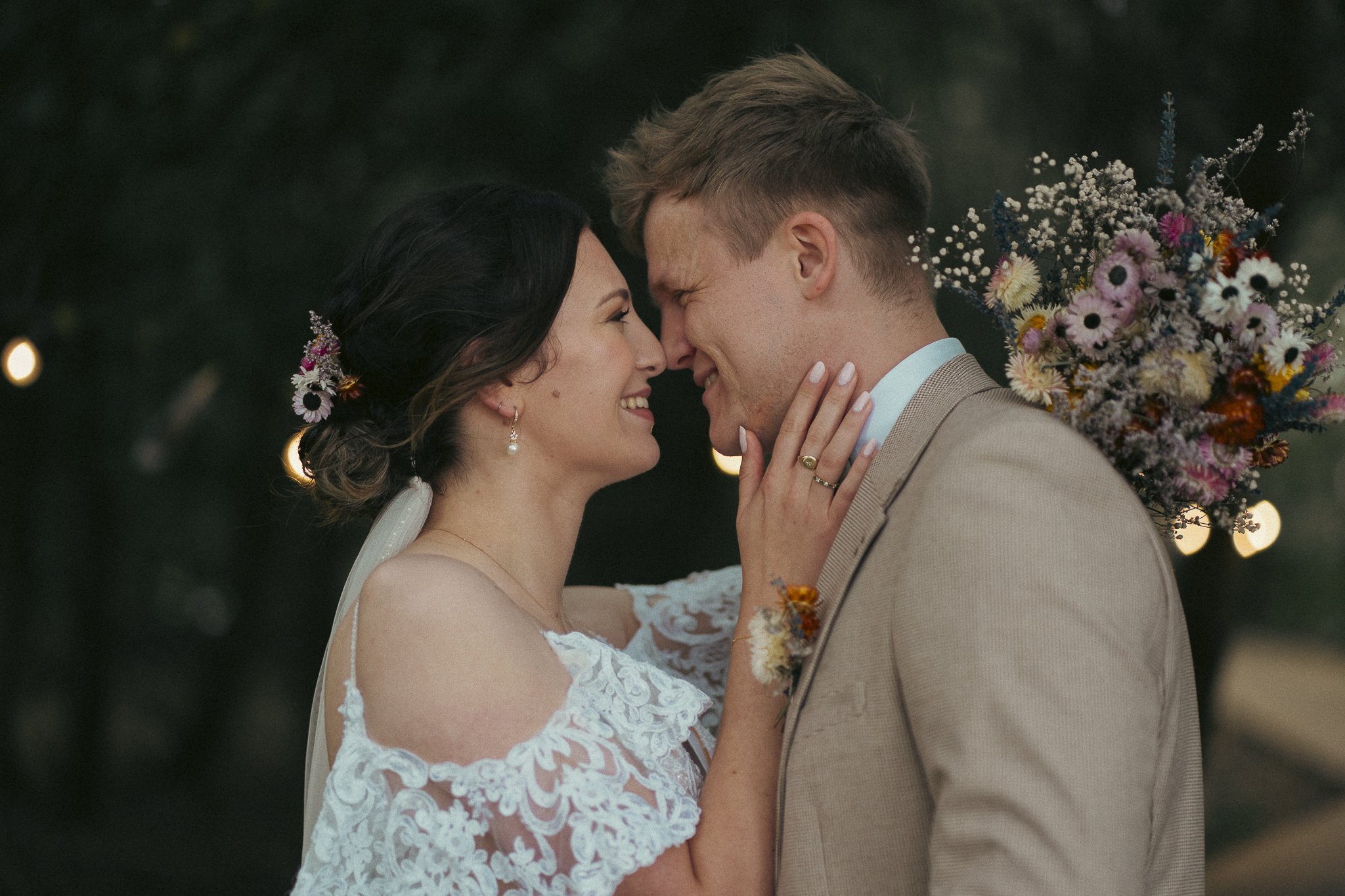 A bride and groom touching foreheads outdoors during wedding ceremony, with the bride holding a bouquet of colorful flowers behind her back.