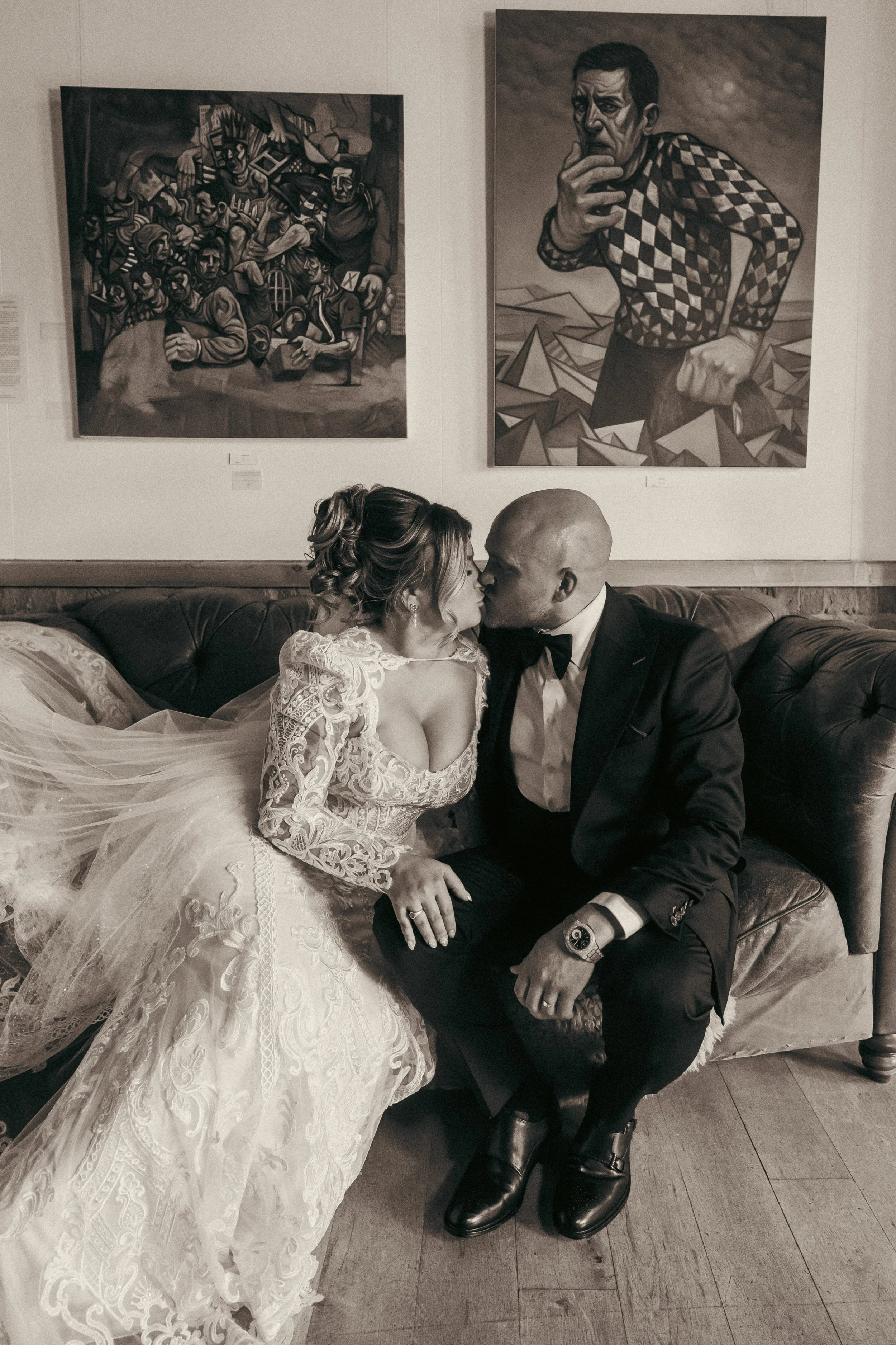 A bride and groom sitting close on a vintage leather sofa, about to kiss, with colorful abstract paintings on the wall behind them.