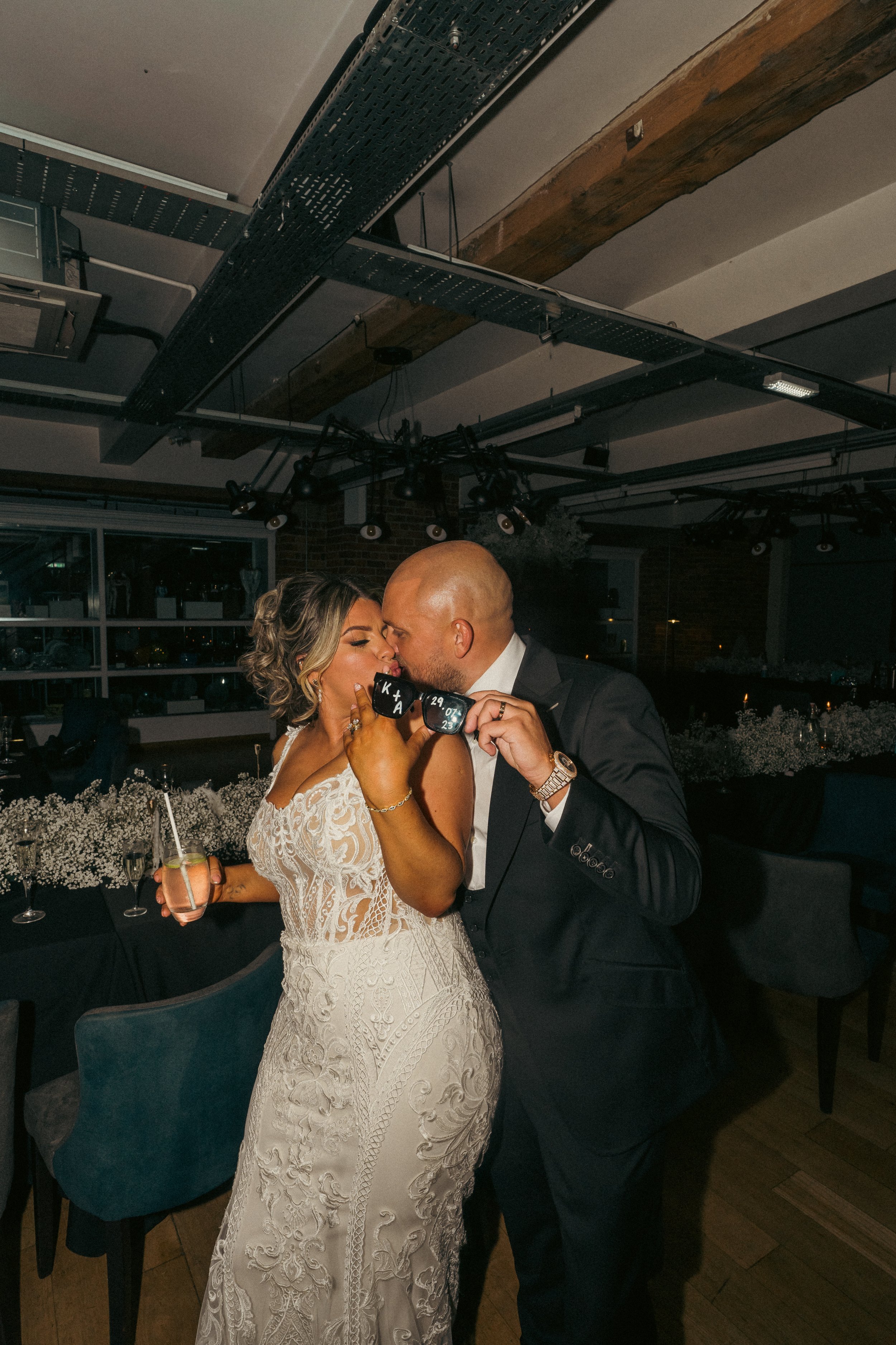 A bride and groom sharing a kiss with sunglasses during their wedding reception in a decorated indoor venue with flowers and tables.