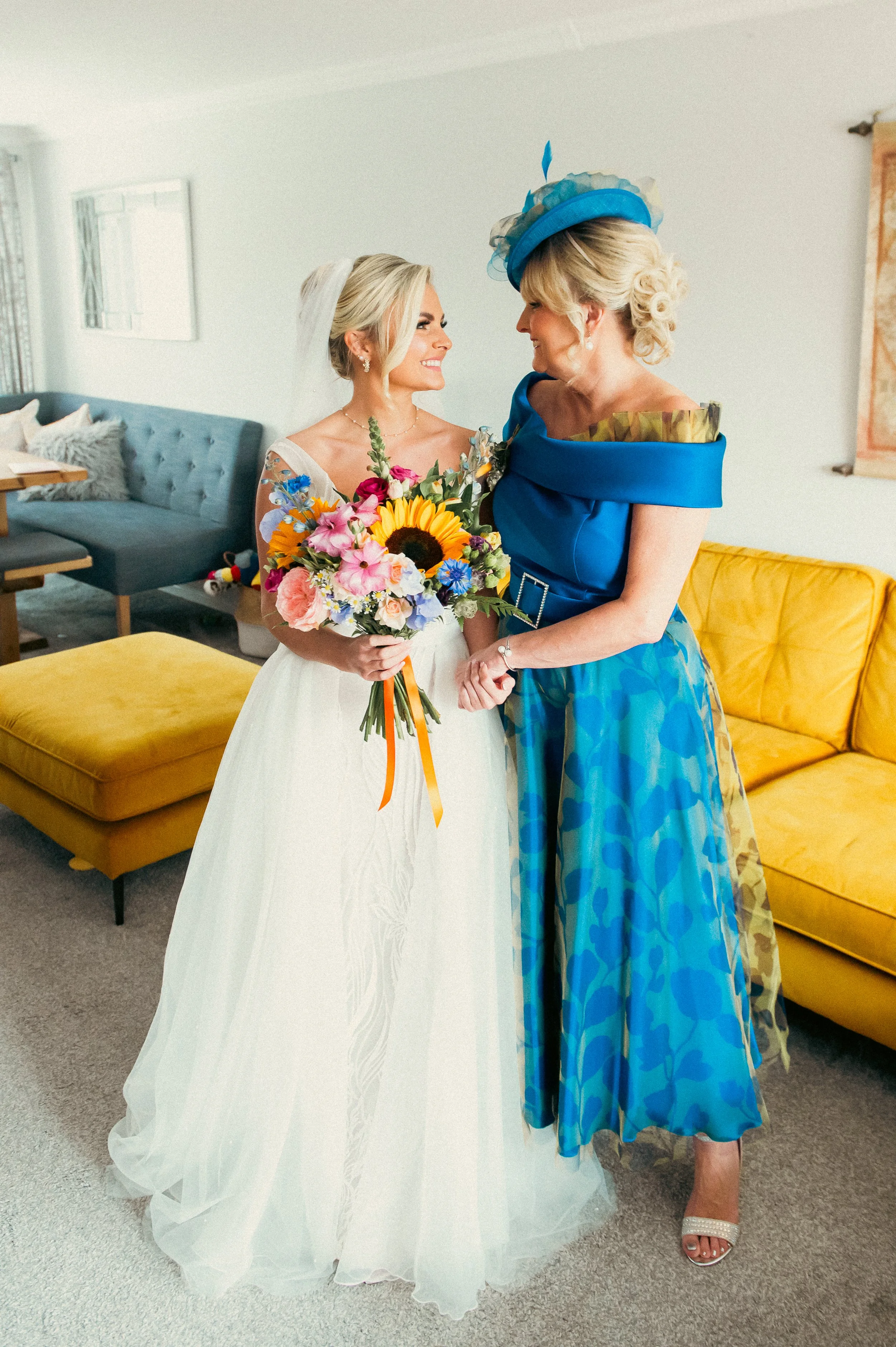 A bride in a white wedding dress holding a colorful bouquet of flowers, smiling at an older woman in a vibrant blue dress and matching hat inside a living room.