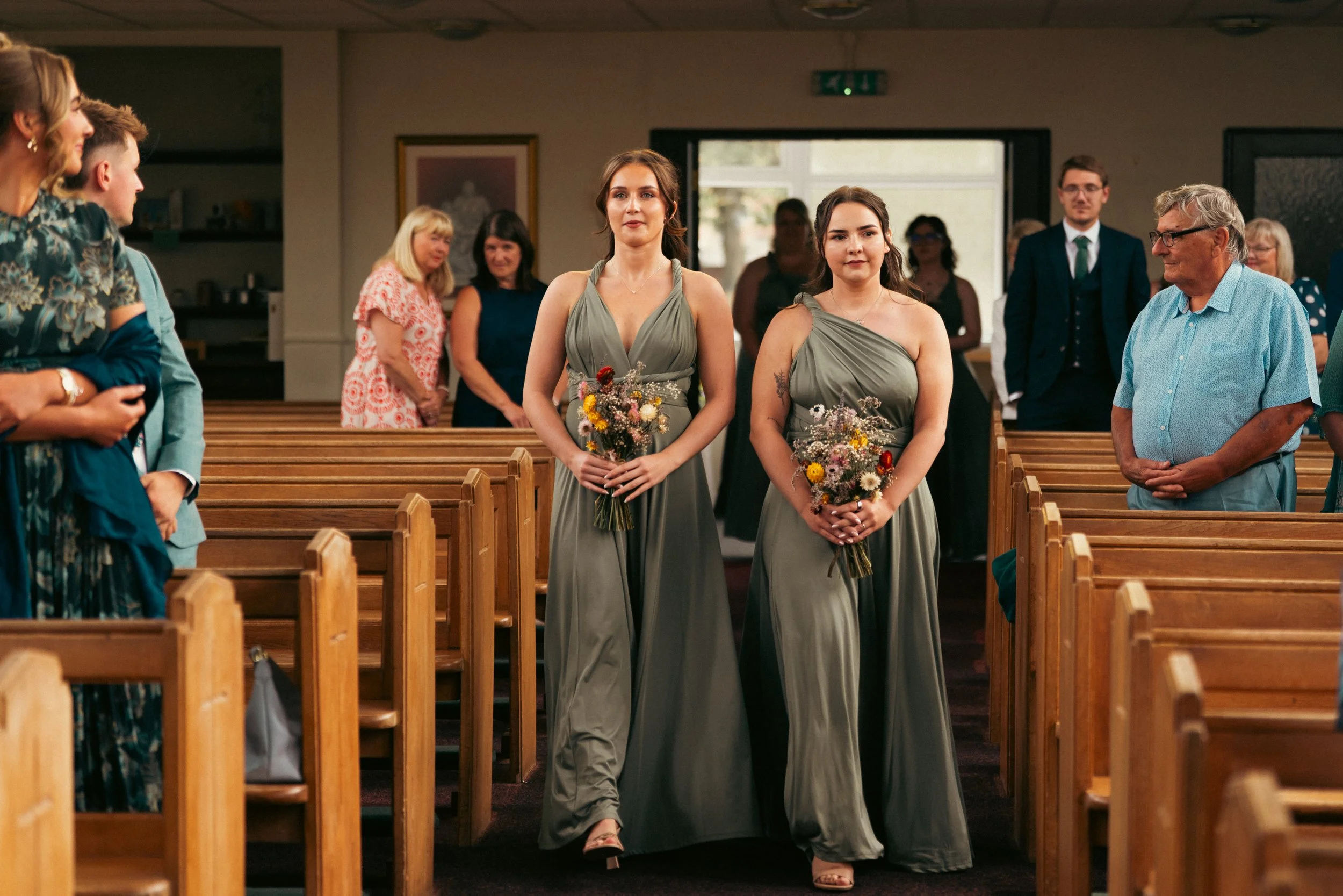 Two women in matching gray dresses holding bouquets walk down the aisle in a church. Guests are standing and watching.