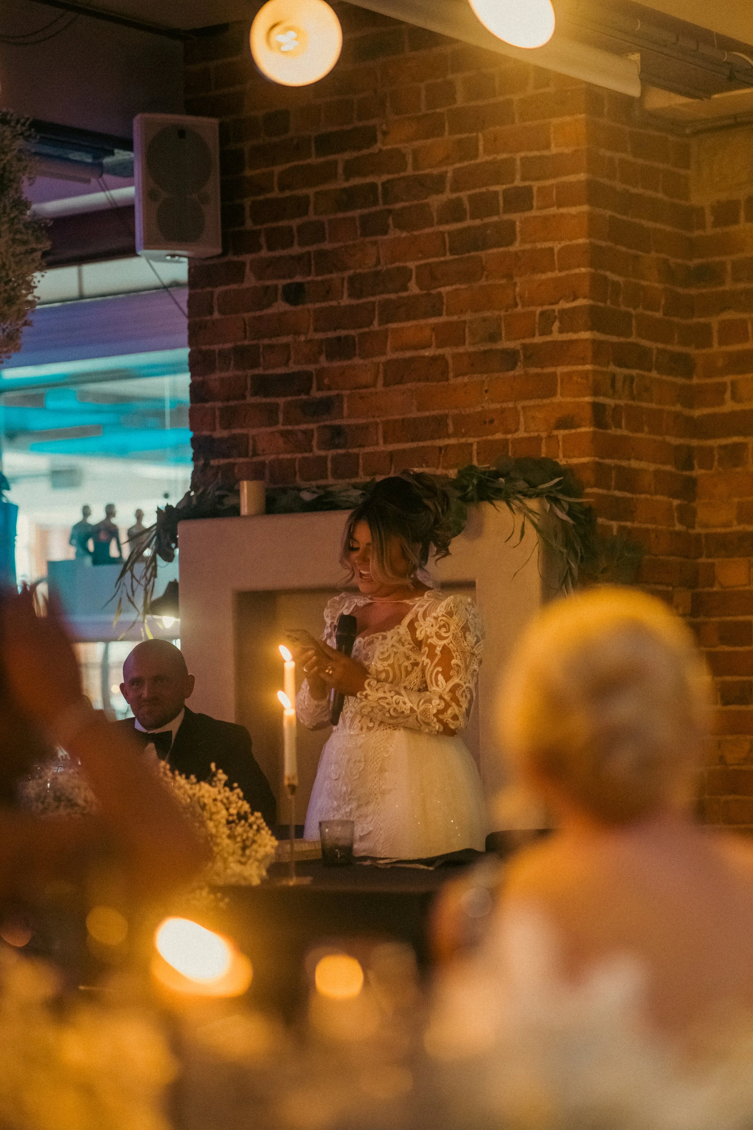 A woman in a lace wedding dress stands at a table holding a microphone and reading from her phone at a wedding reception. There are candles and floral arrangements on the table, a brick wall in the background, and a man in a tuxedo sitting nearby.