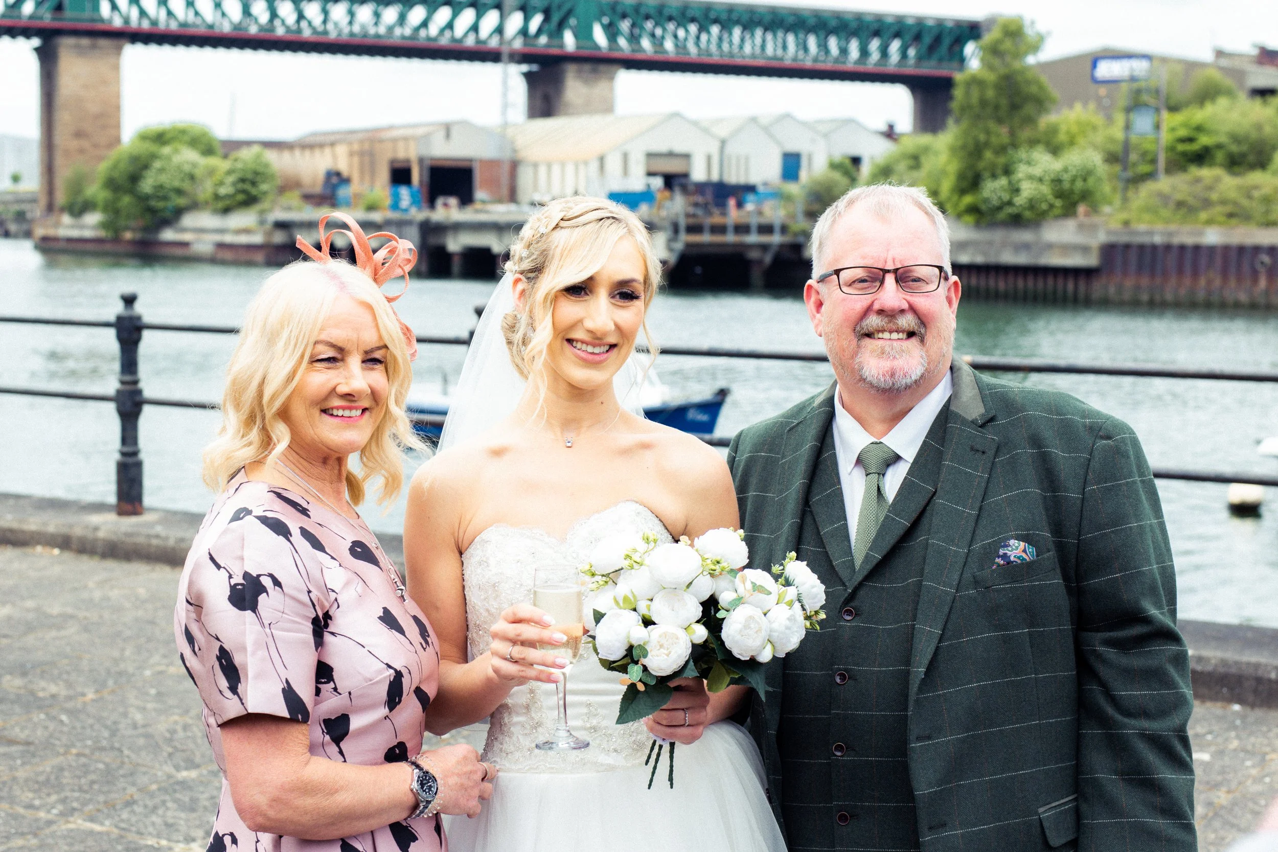 A bride in a white wedding dress holding a bouquet of white flowers and a glass of champagne, standing next to a woman in a pink and black dress with a fascinator, and a man in a green checkered suit with glasses, all smiling near the water with boat