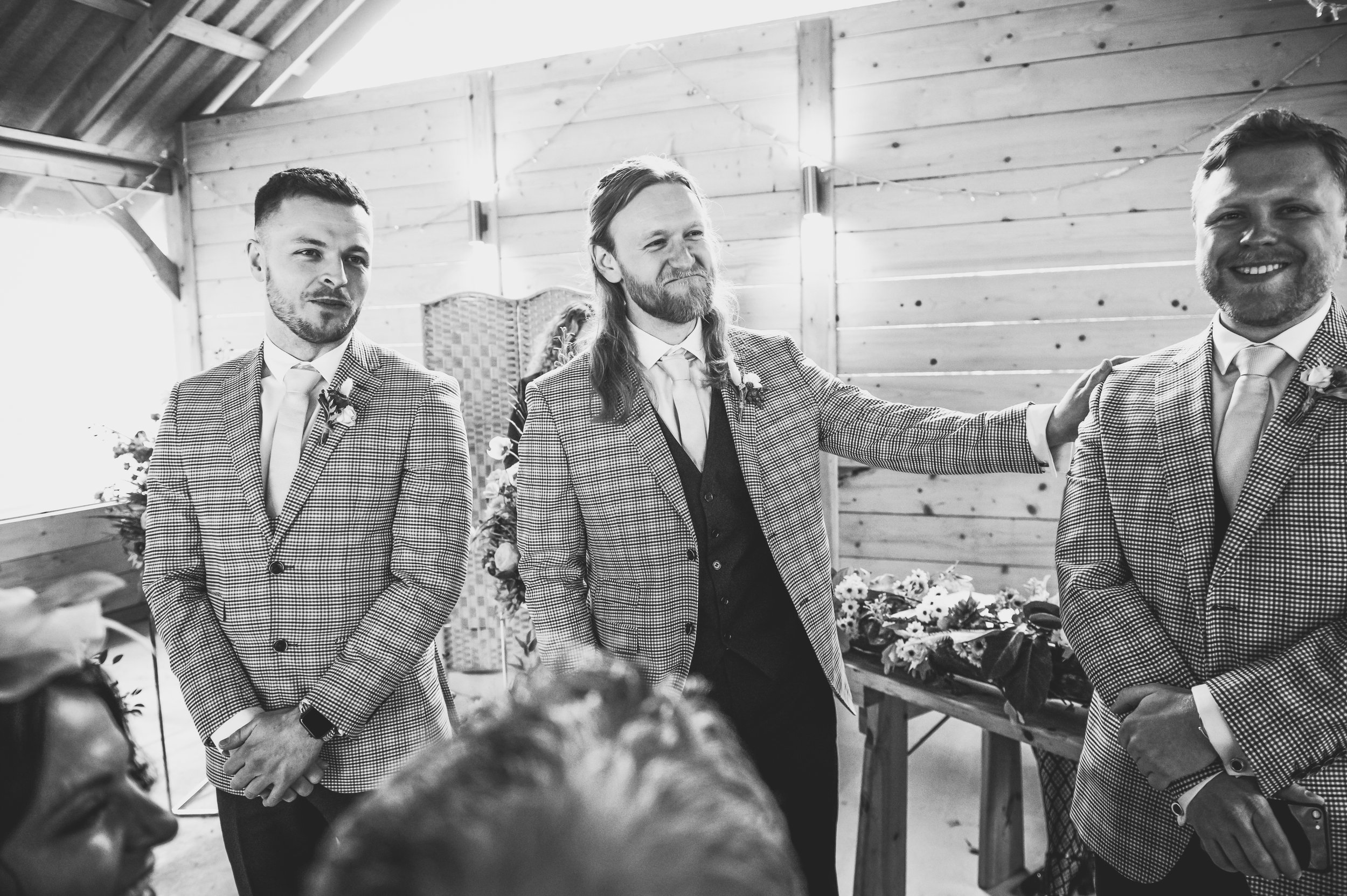 Black-and-white photo of three men holding a wedding vow exchange or wedding ceremony, with one in the middle smiling and touching the shoulder of the man on the right, inside a wooden building decorated with flowers.