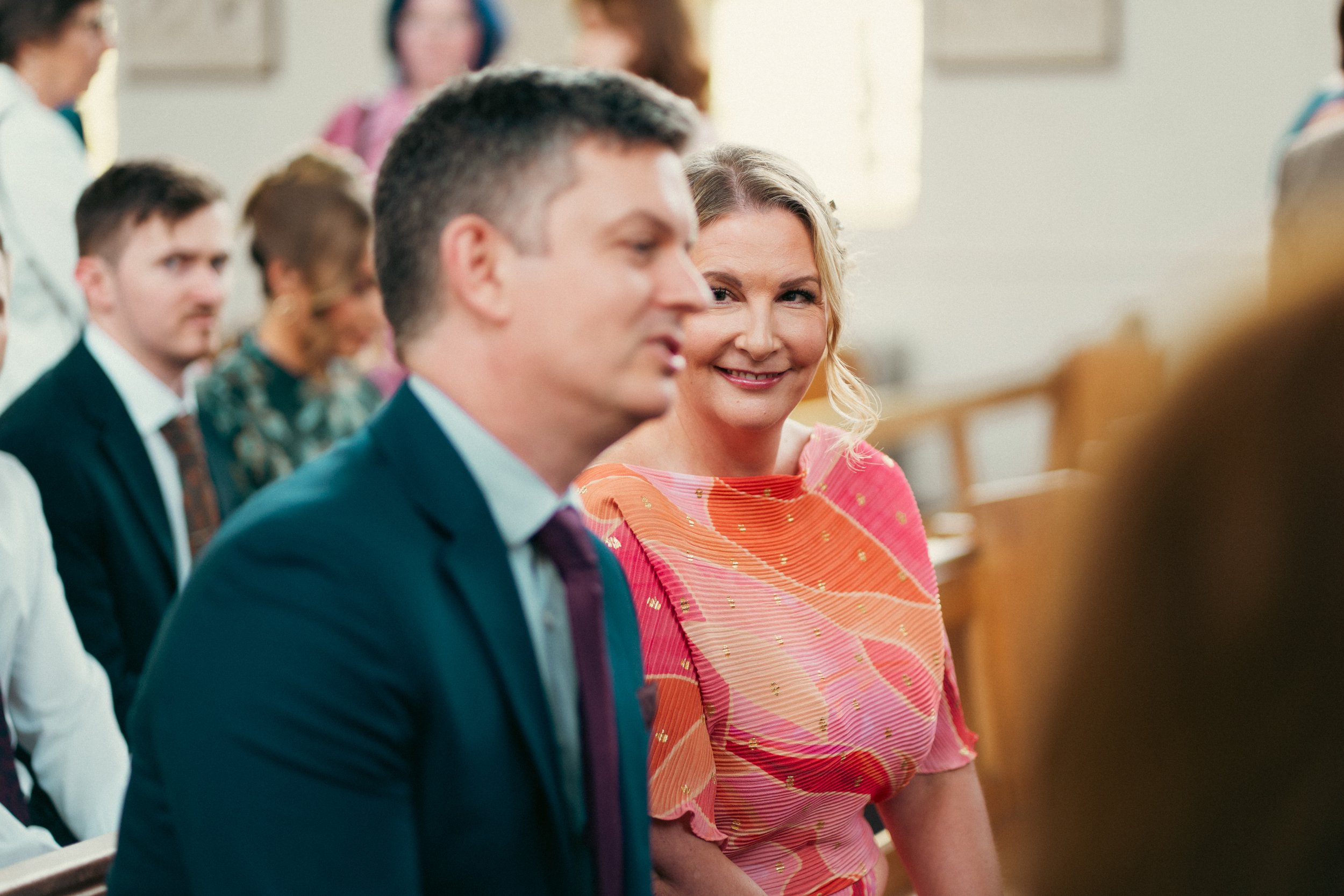 A woman in a pink and orange dress smiling at a man in a blue suit during a church ceremony, with other people sitting in the background.