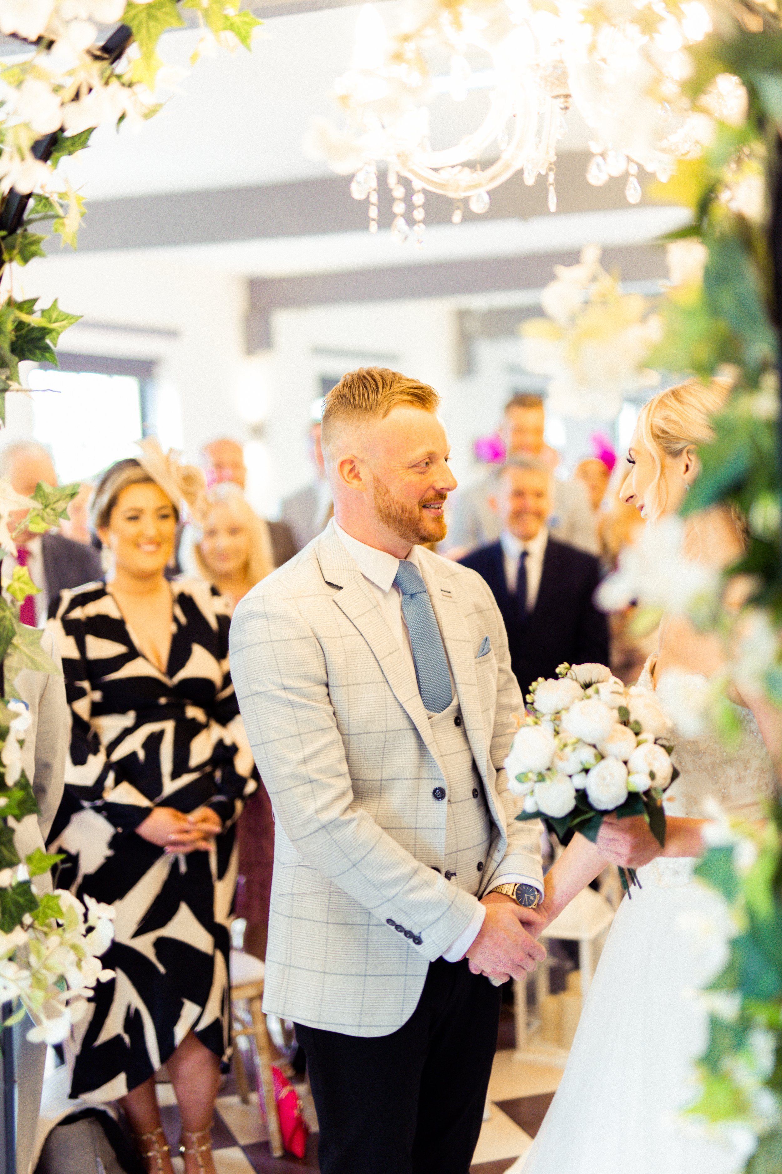 A groom standing and holding a bouquet of white flowers during a wedding ceremony, with guests smiling in the background.