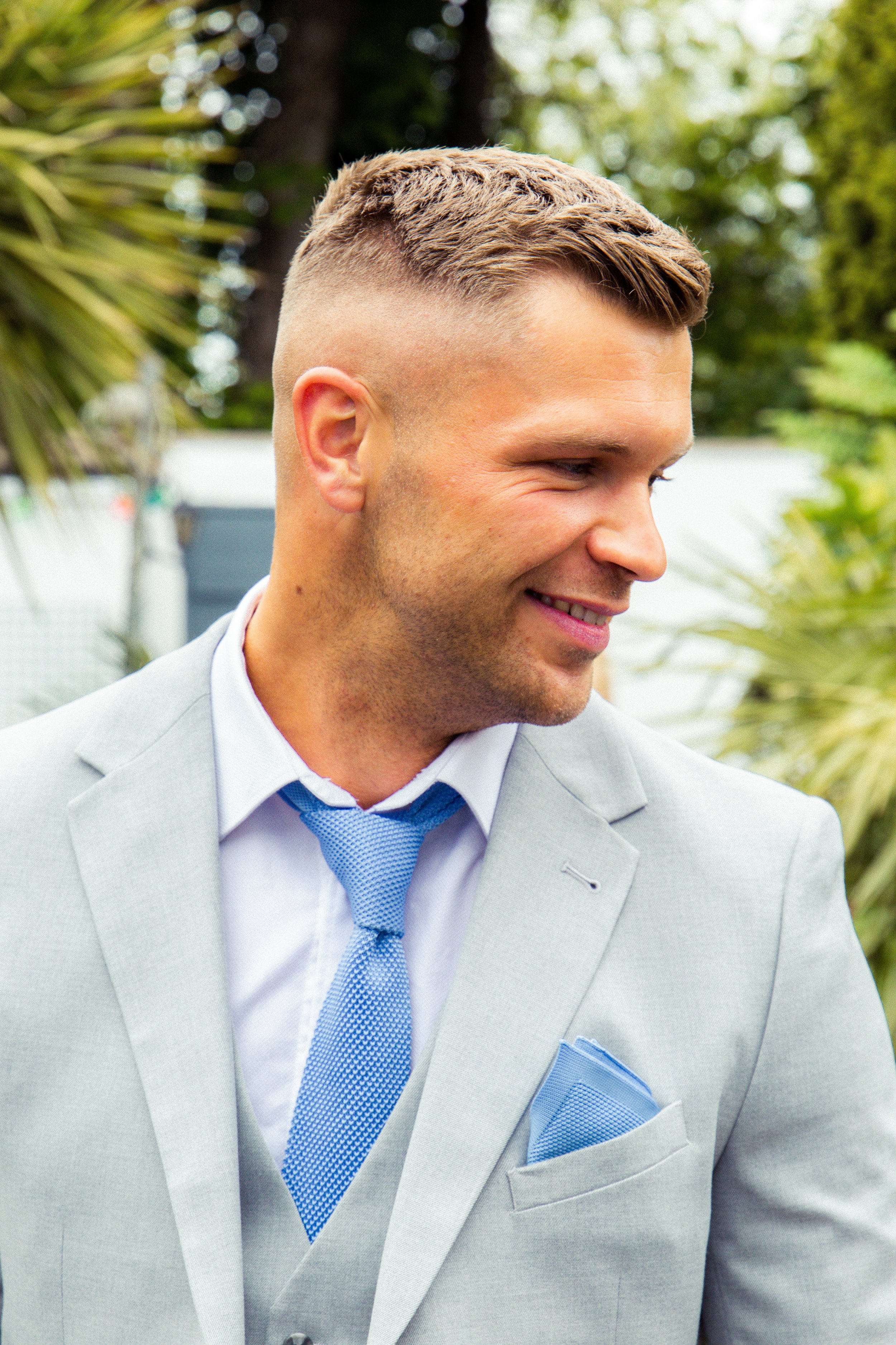 Smiling man in a light gray suit with a blue tie and pocket square, outdoors with greenery in the background.