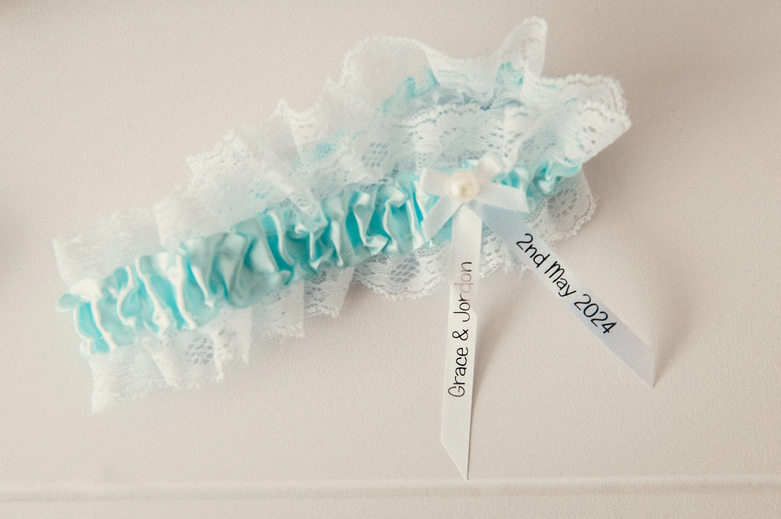 Light blue and white lace wedding garter with bows and pearls, with attached satin ribbons inscribed with "Grace & Jordan" and the date "2nd May 2024".