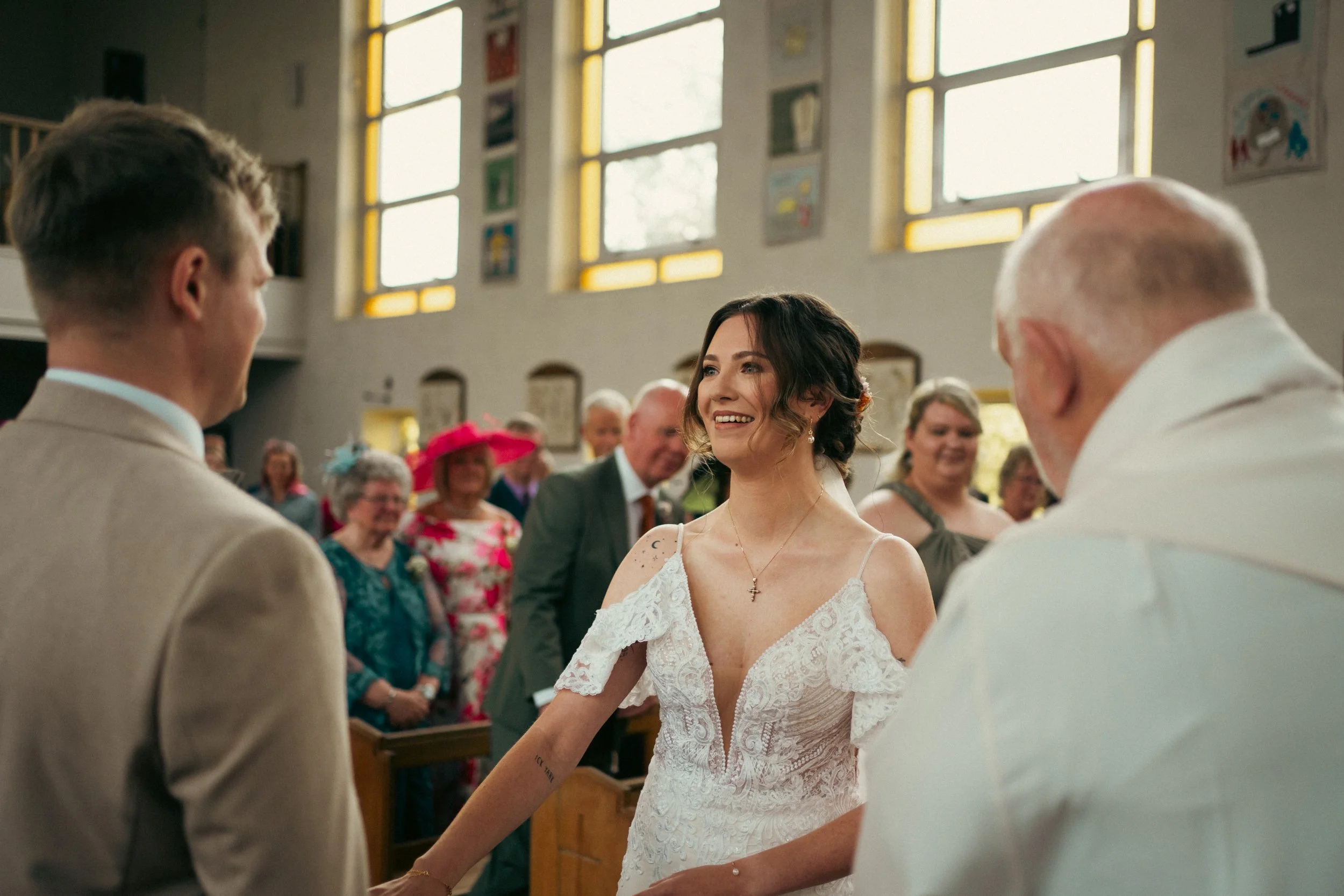 A woman in a white wedding dress smiling at a man in a beige suit during a wedding ceremony inside a church with stained glass windows.