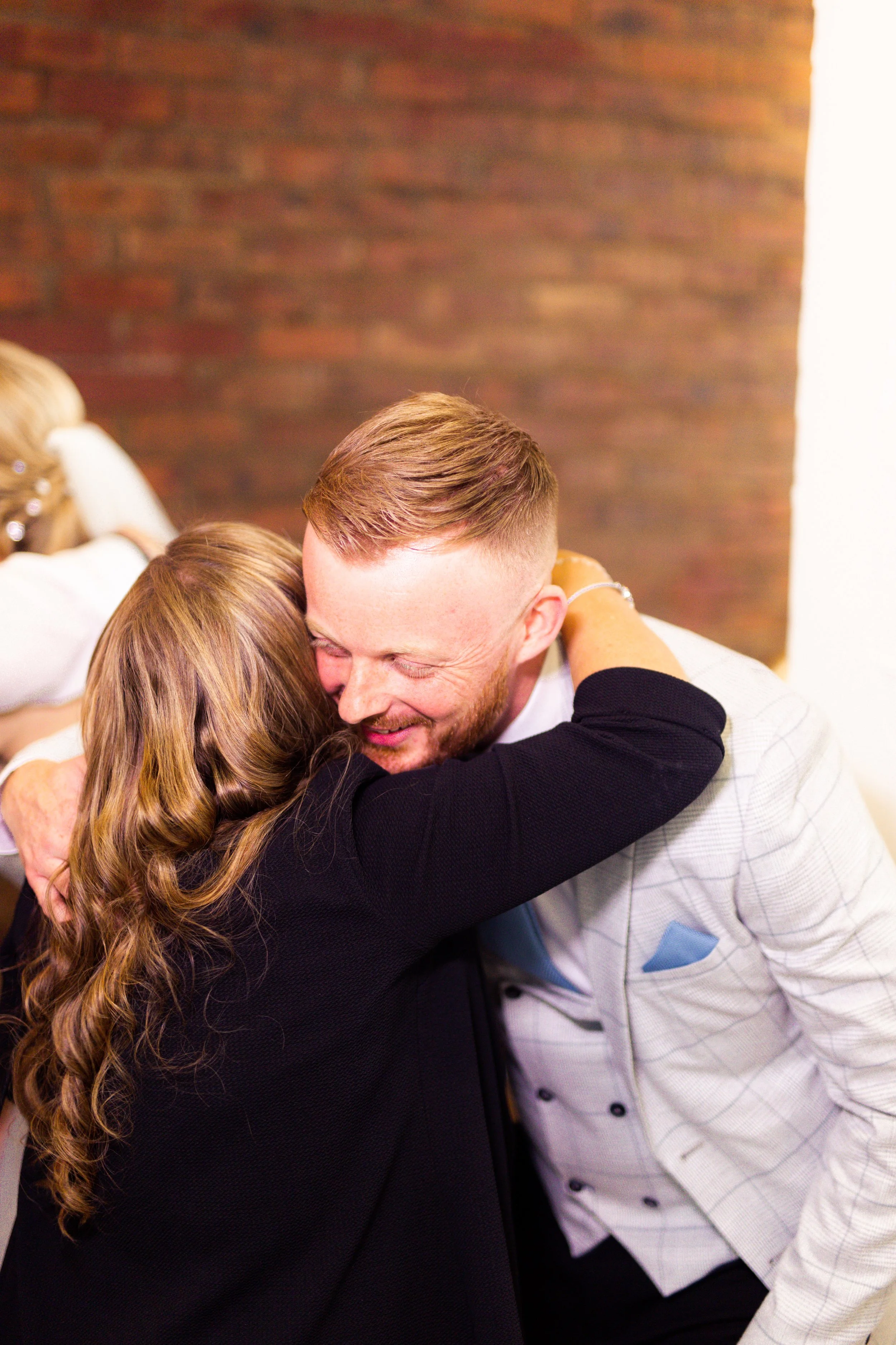 A man and a woman warmly hugging at a social event, with other people in the background.