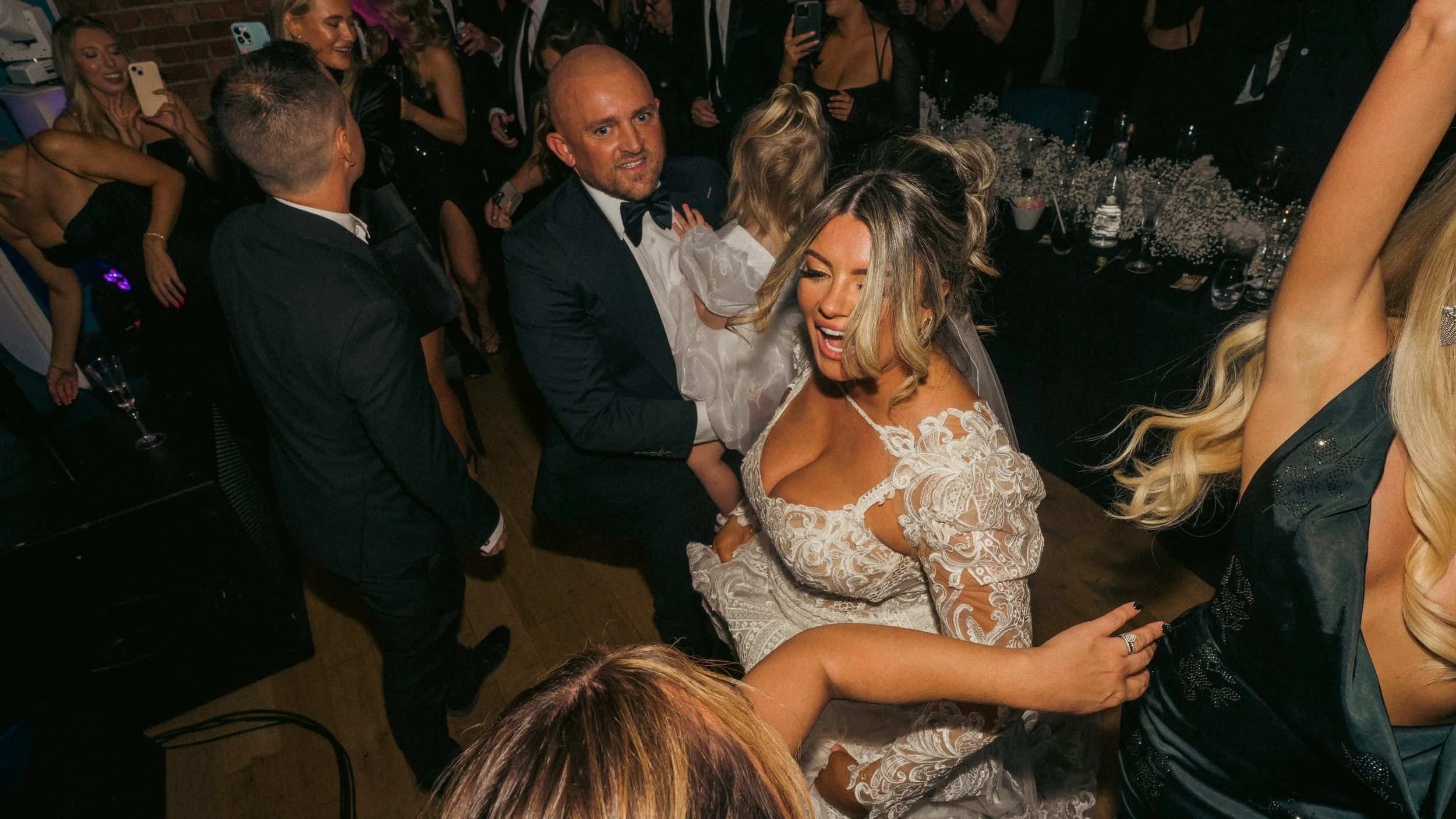 Group of people dancing and celebrating at a party, including a woman in a white lace dress and a man in a black tuxedo holding a girl in a white dress, with others in formal attire around them.