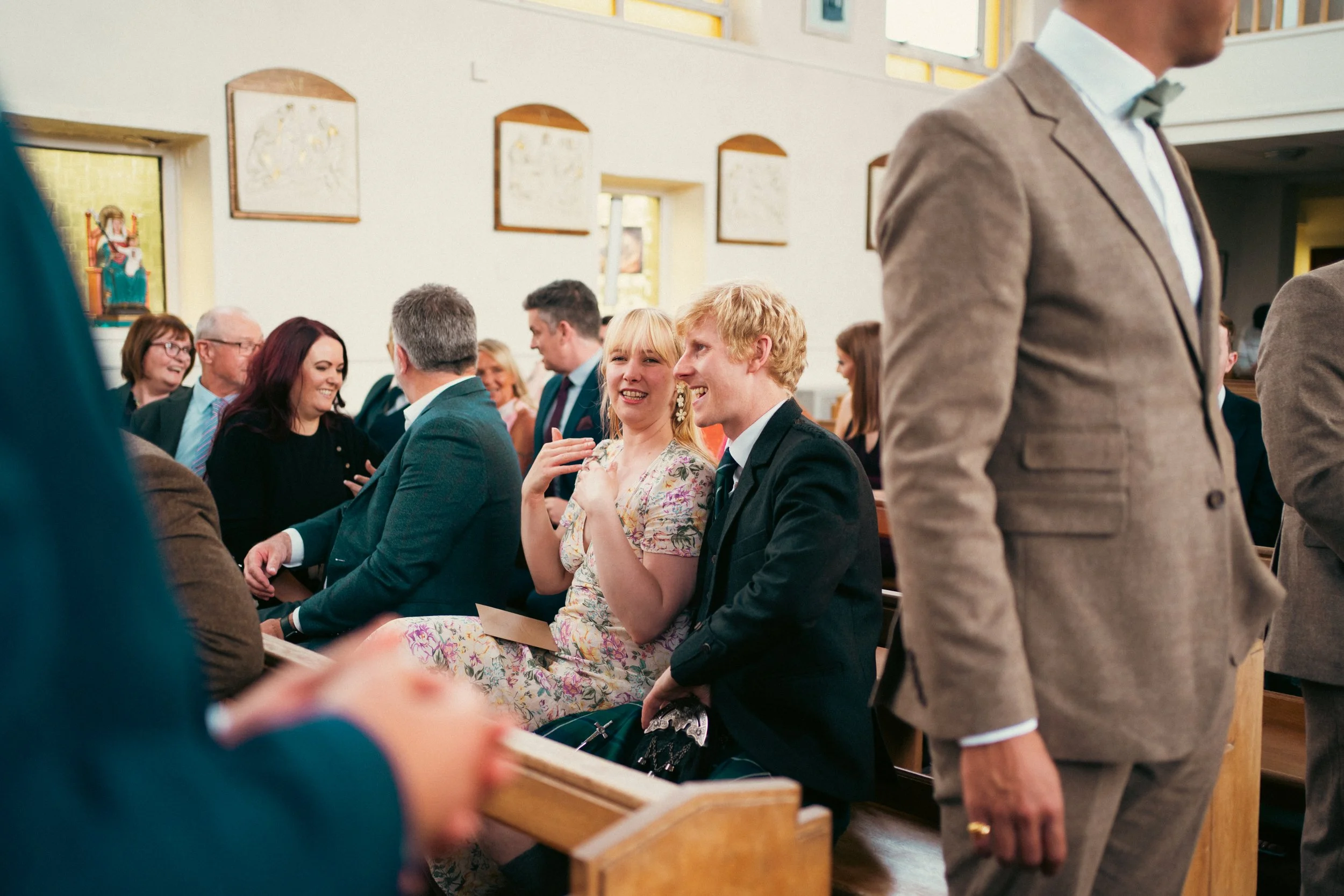 People sitting and standing in a church during a wedding ceremony, smiling and engaging with each other.