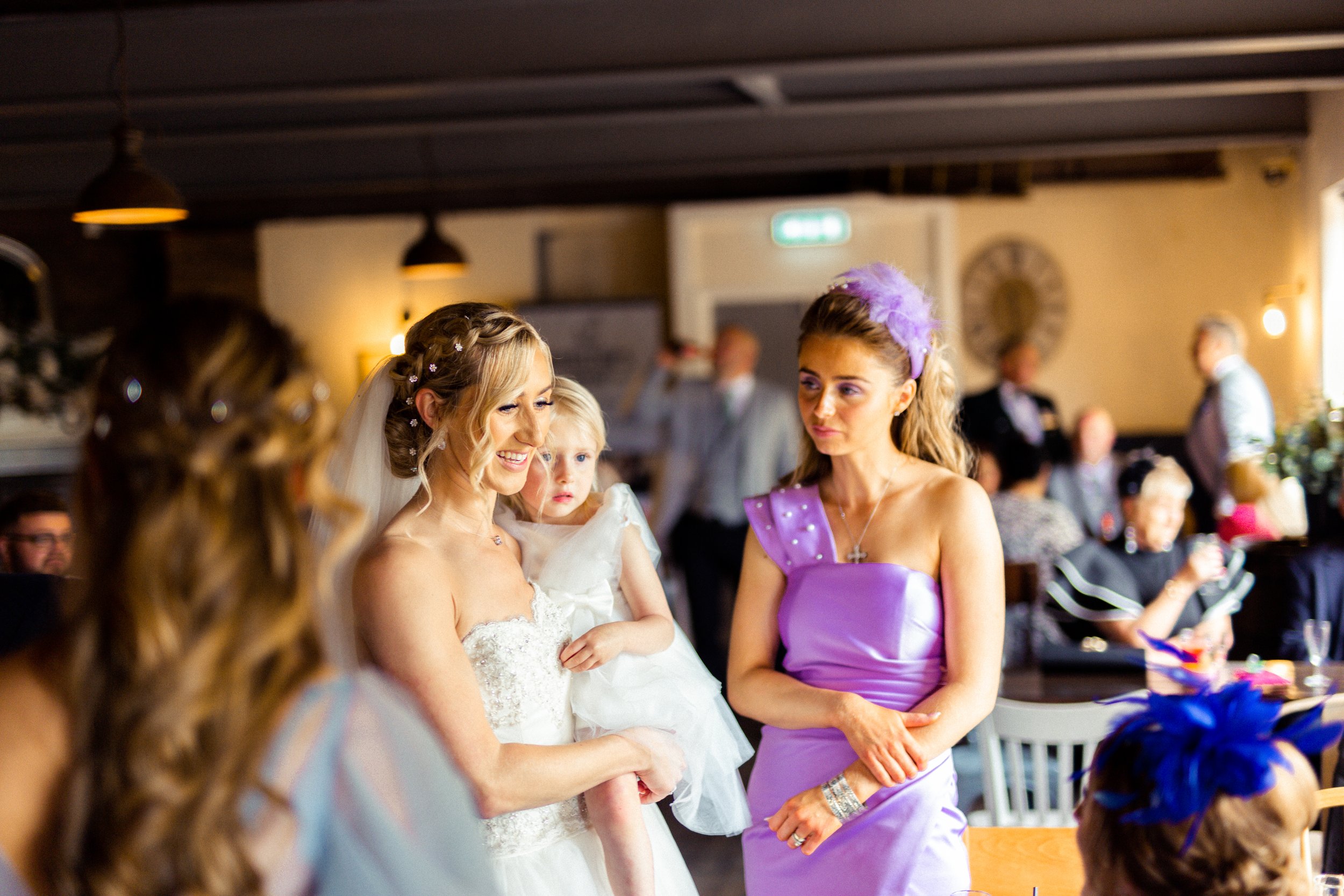 A bride in a white wedding dress holding a young girl in a white dress, smiling. Two women are talking to her—one in a purple dress with a feathered headband, and another partially visible with curly hair and a floral headband. The background shows p
