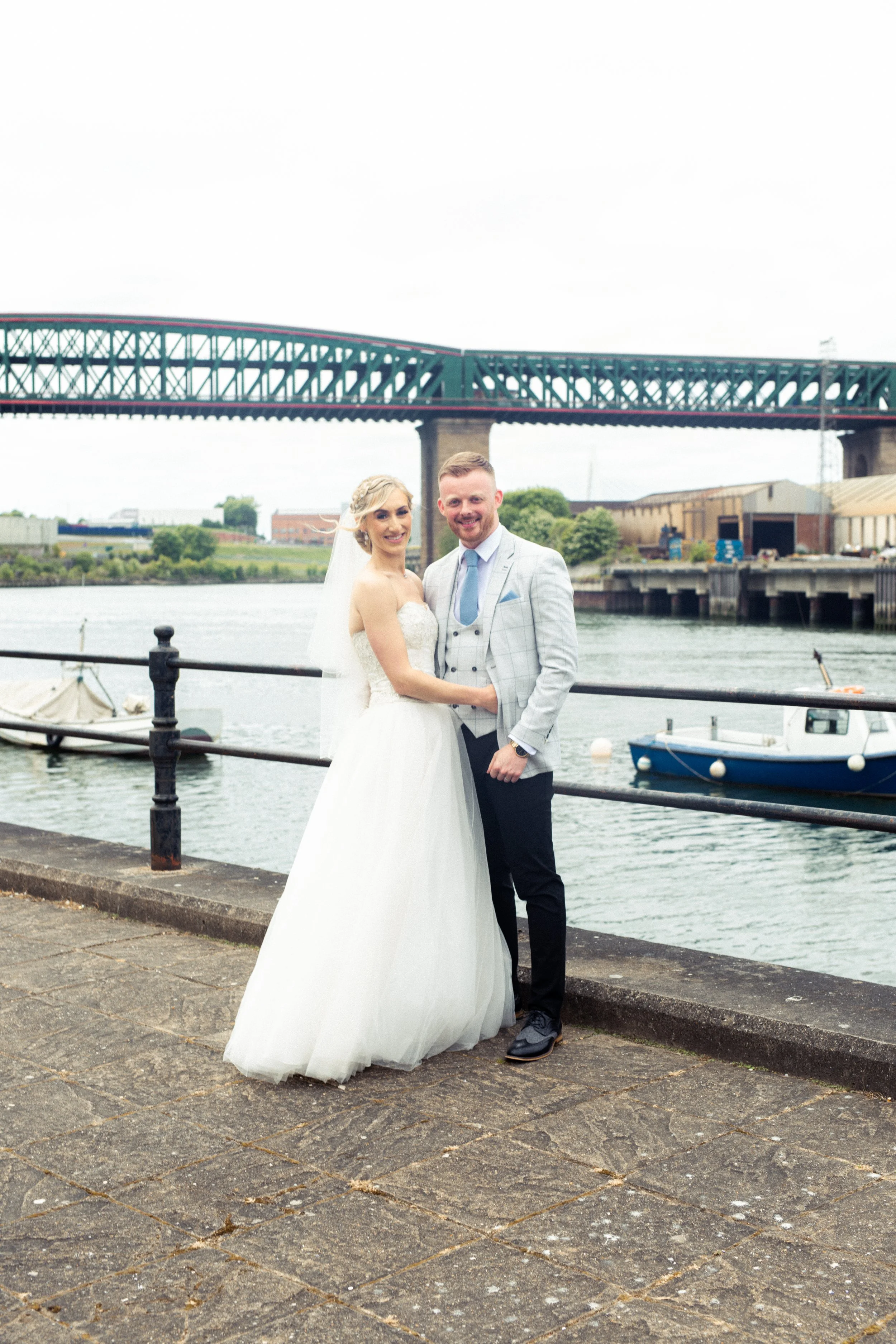 A newlywed couple stands arm-in-arm on a riverside promenade with boats and a bridge in the background. The bride wears a white wedding gown and veil, and the groom wears a light gray checkered suit with a blue tie.