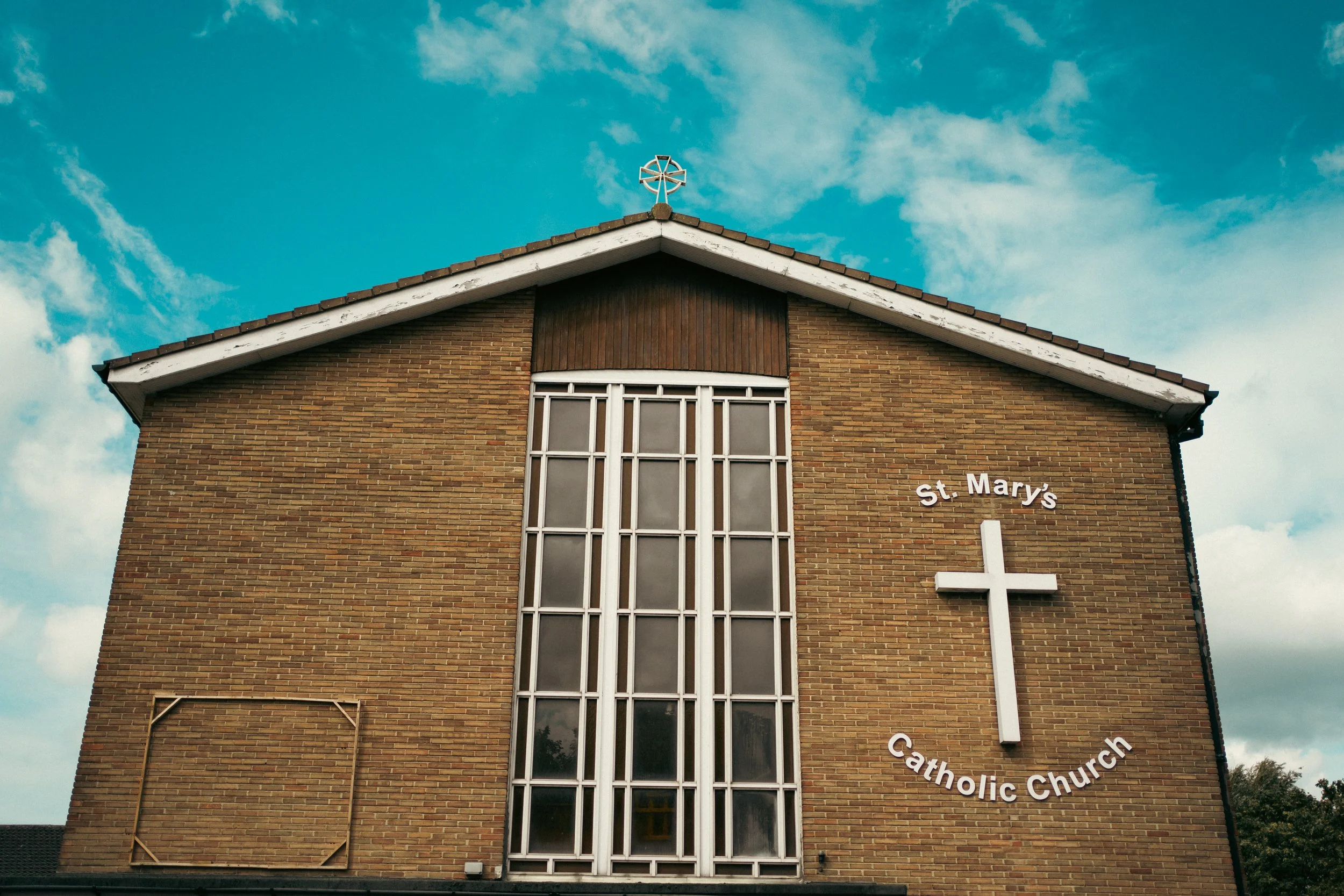 Front view of St. Mary's Catholic Church building with brick exterior, large window, and white cross on the wall, under a blue sky with scattered clouds.